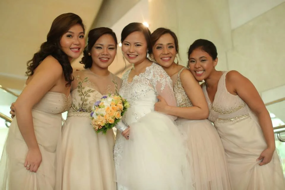 A bride in a white wedding gown surrounded by five bridesmaids in coordinated beige dresses, all smiling indoors.