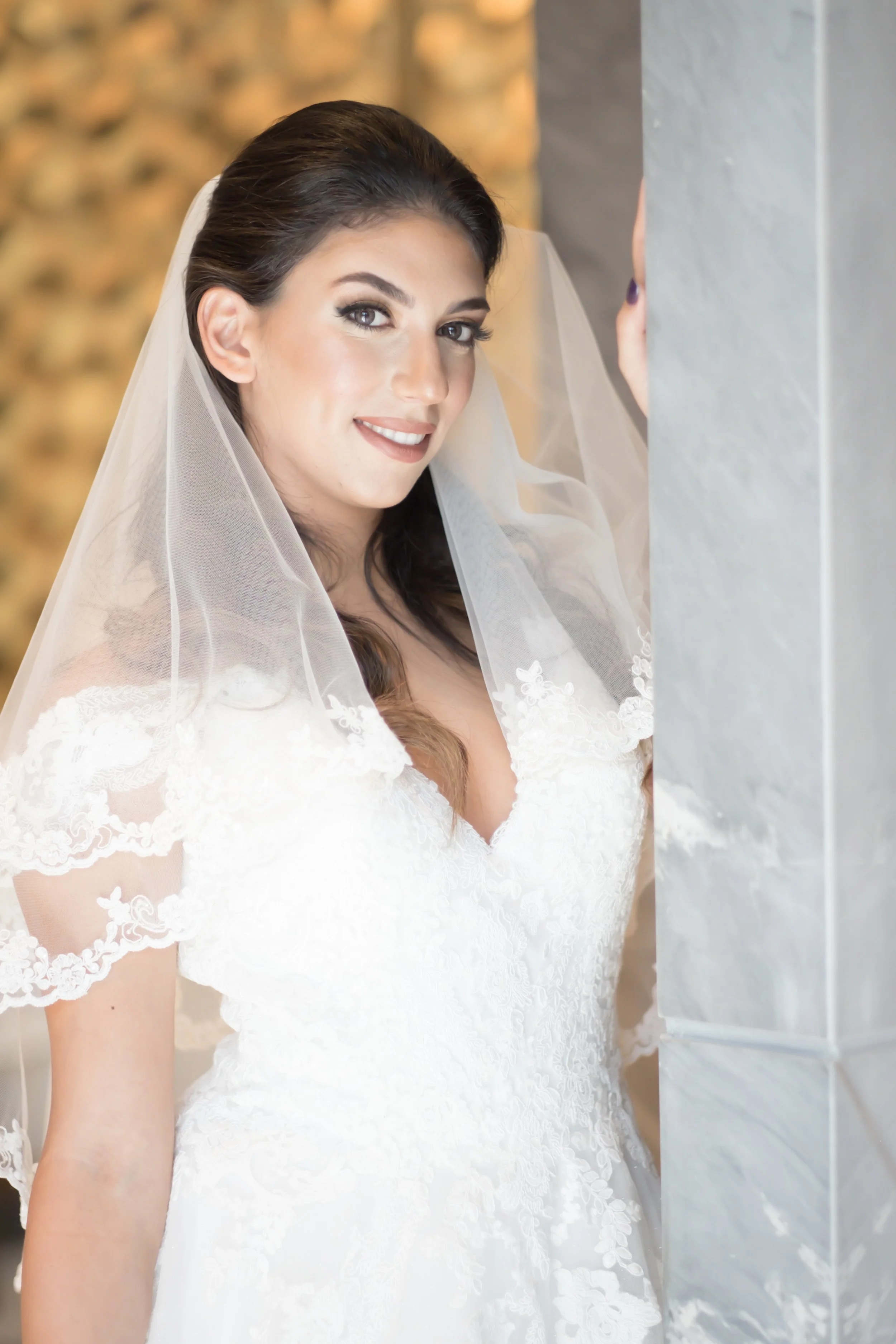 A bride in a white lace wedding gown with a veil, smiling and standing next to a gray stone column.