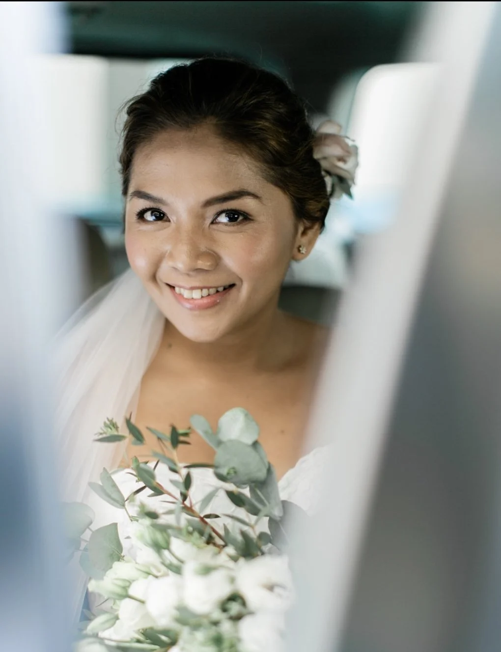 Smiling woman with dark hair in bun, holding a bouquet of white flowers and greenery, looking at the camera.