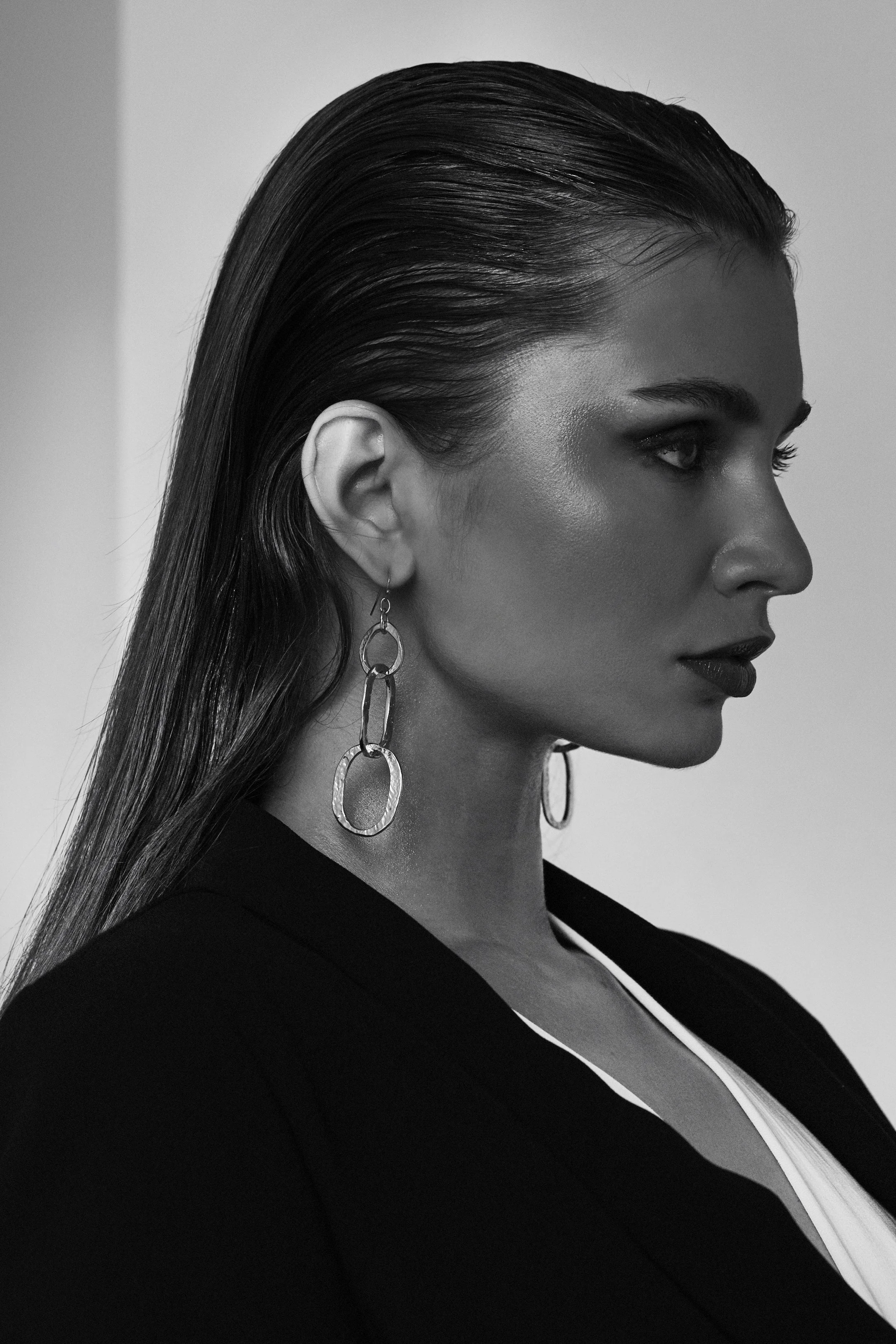 A black and white profile portrait of a woman with sleek, wet-look hair, wearing large linked hoop earrings, a dark blazer, and a white top.