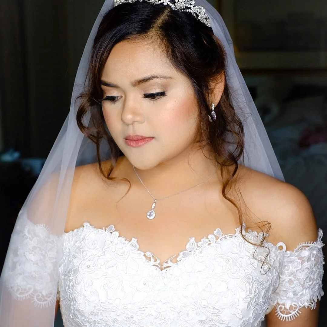 A bride in a white lace wedding dress with a sheer veil and jewelry, looking down with a calm expression.