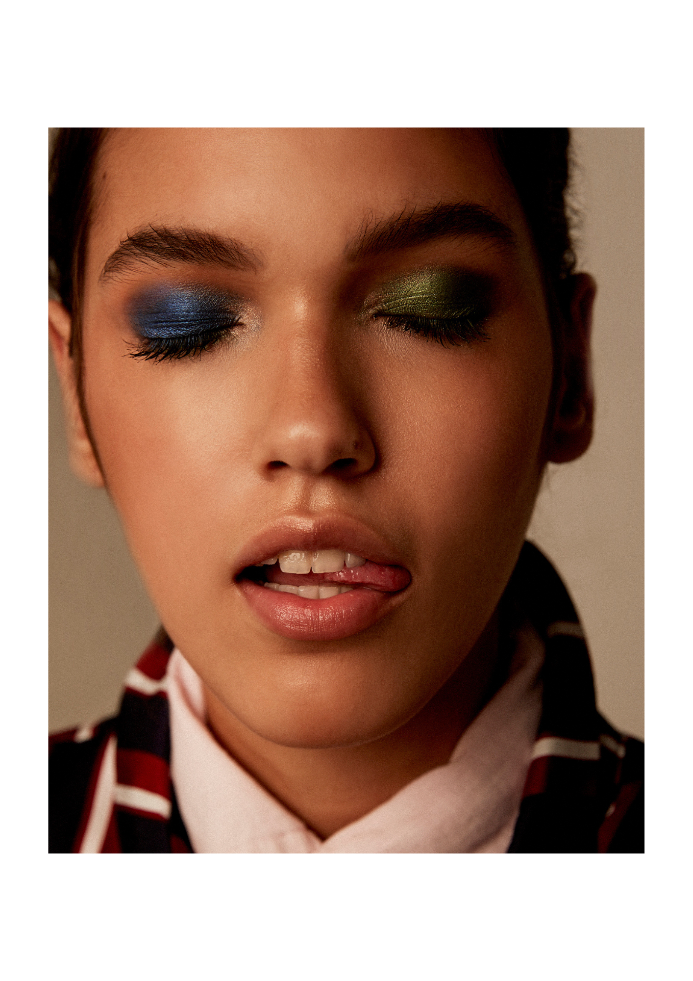 Close-up of a young woman with colorful makeup, eyes closed, tongue slightly out and lips parted, wearing a patterned shirt and a white scarf.
