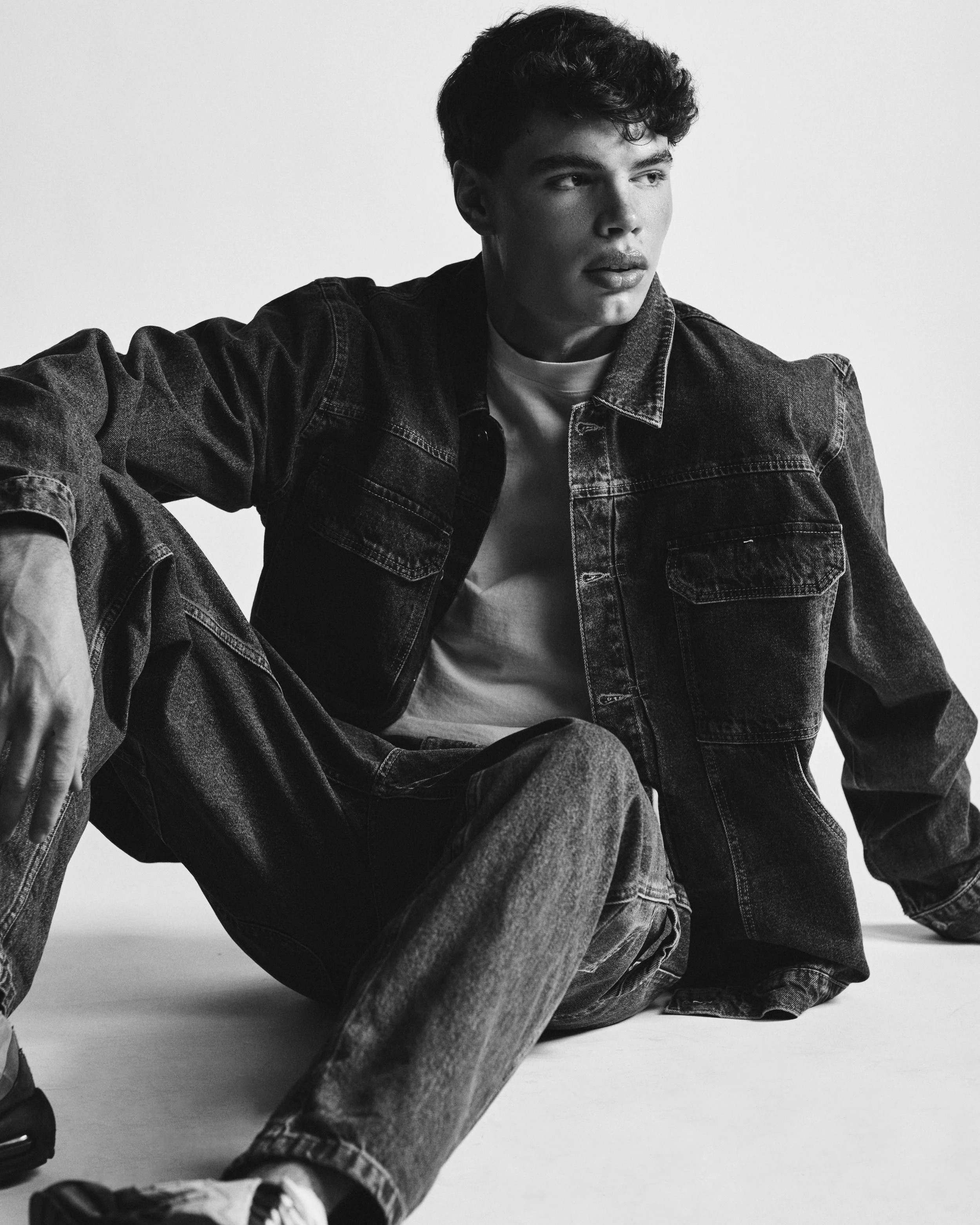 Black and white photo of a young man in a denim jacket and jeans, sitting on the floor and looking to the side.
