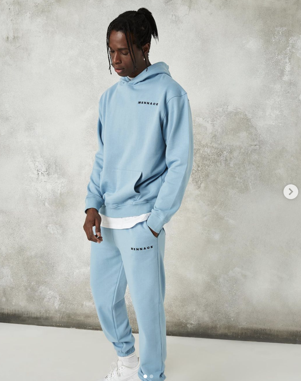 A man wearing a light blue hoodie and matching sweatpants with the word 'MENNACE' printed on both, standing against a textured gray wall, with his head tilted downward.