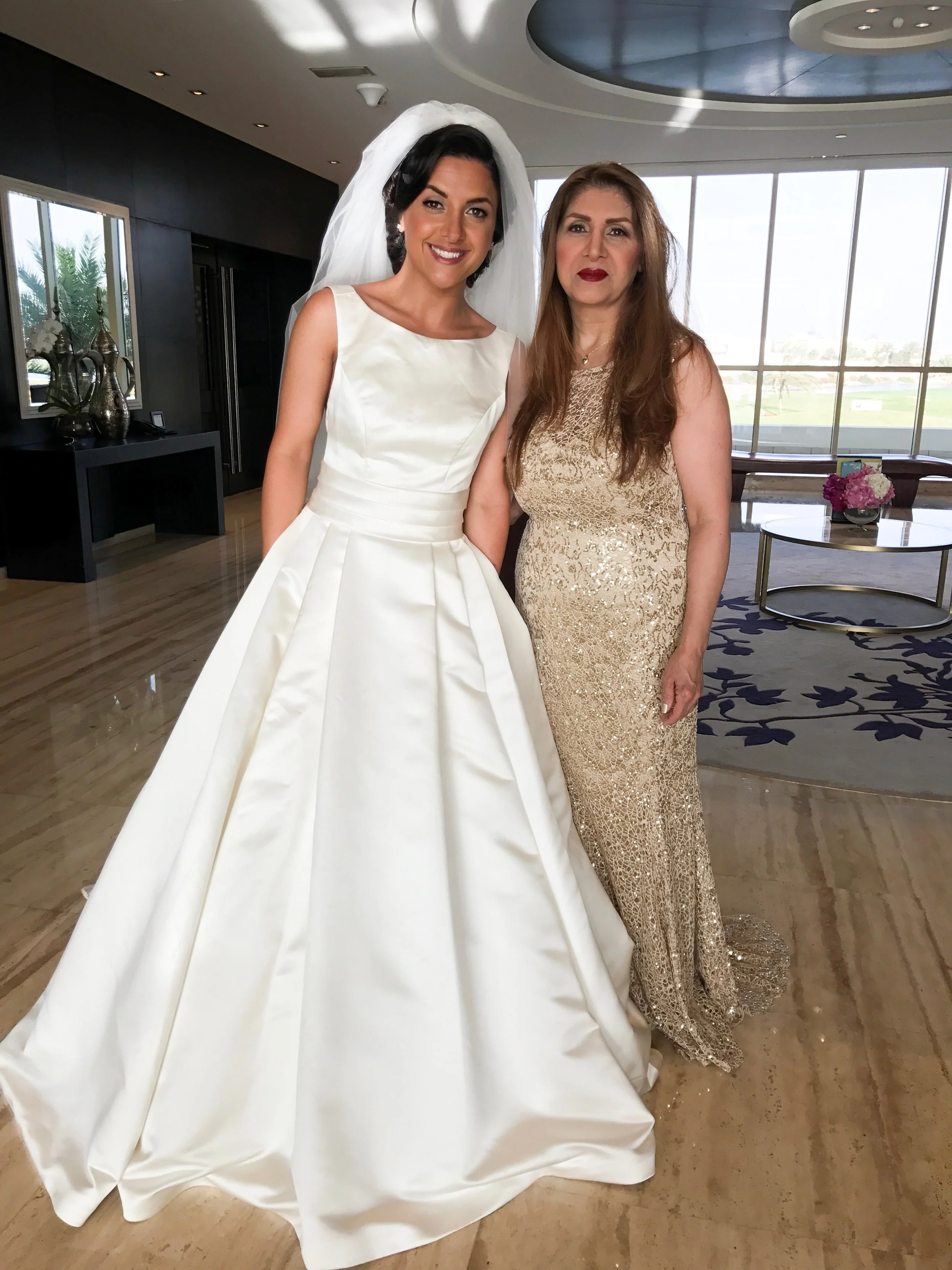 A bride in a white wedding dress and veil standing next to an older woman in a gold gown inside a modern room with large windows and a scenic view.