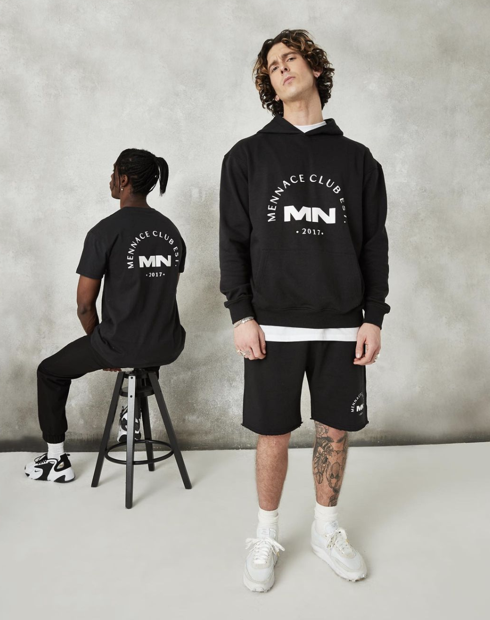 Two young men wearing black men’s athletic clothing with 'MENNACE CLUB EST.' logos, one standing and the other sitting on a stool in front of a gray textured wall.