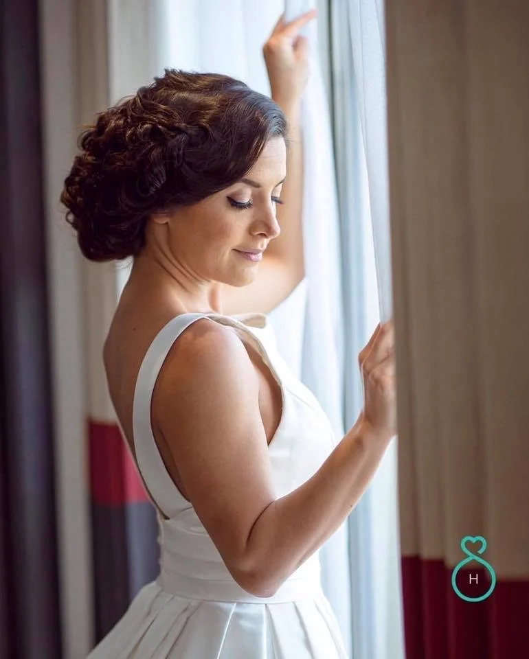 A woman with short, curly brown hair standing by a window, looking down with a gentle smile, wearing a white sleeveless dress.