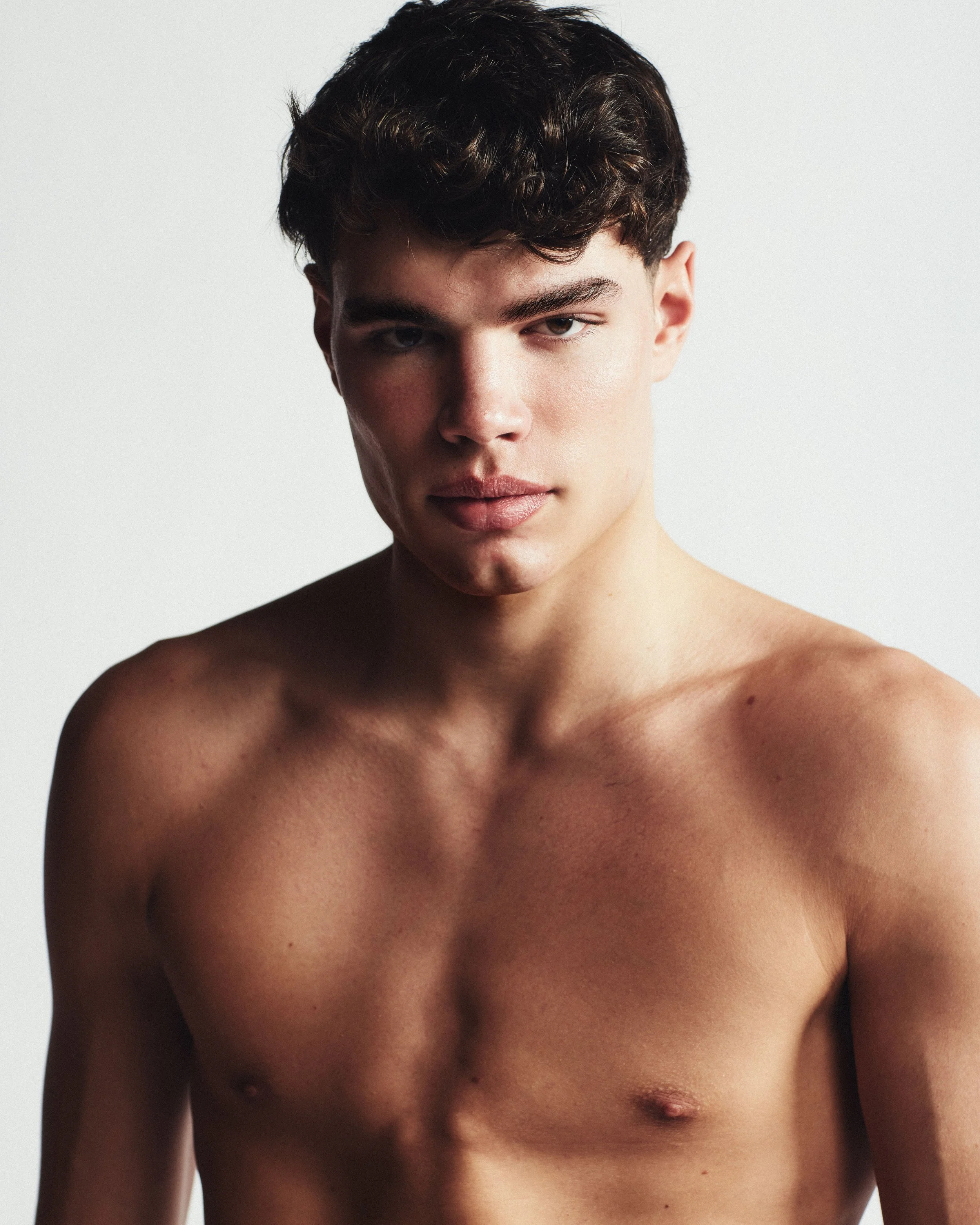 Portrait of a young man with short dark curly hair, shirtless, with a neutral expression, standing against a plain light background.