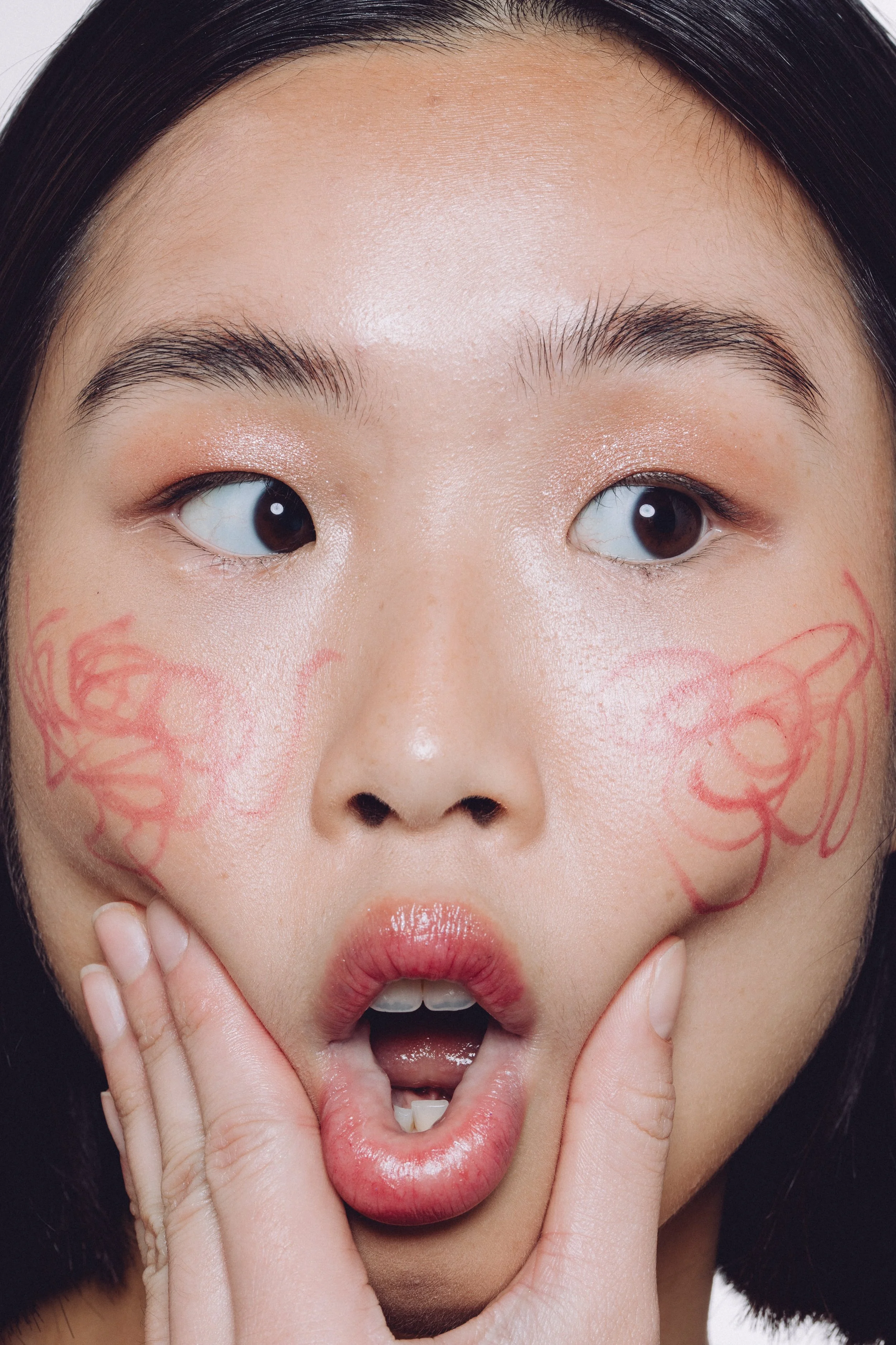 A close-up of a woman with painted red designs on her cheeks, holding her chin with her fingers, with her mouth open and eyes looking to the side.
