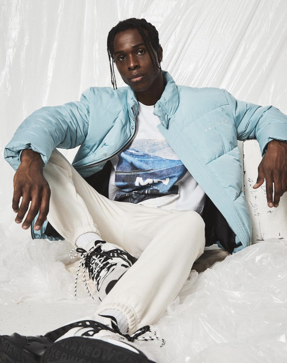 A young man with dreadlocks wearing a light blue puffer jacket, graphic T-shirt, white joggers, and sneakers, sitting against a white background with plastic sheets.