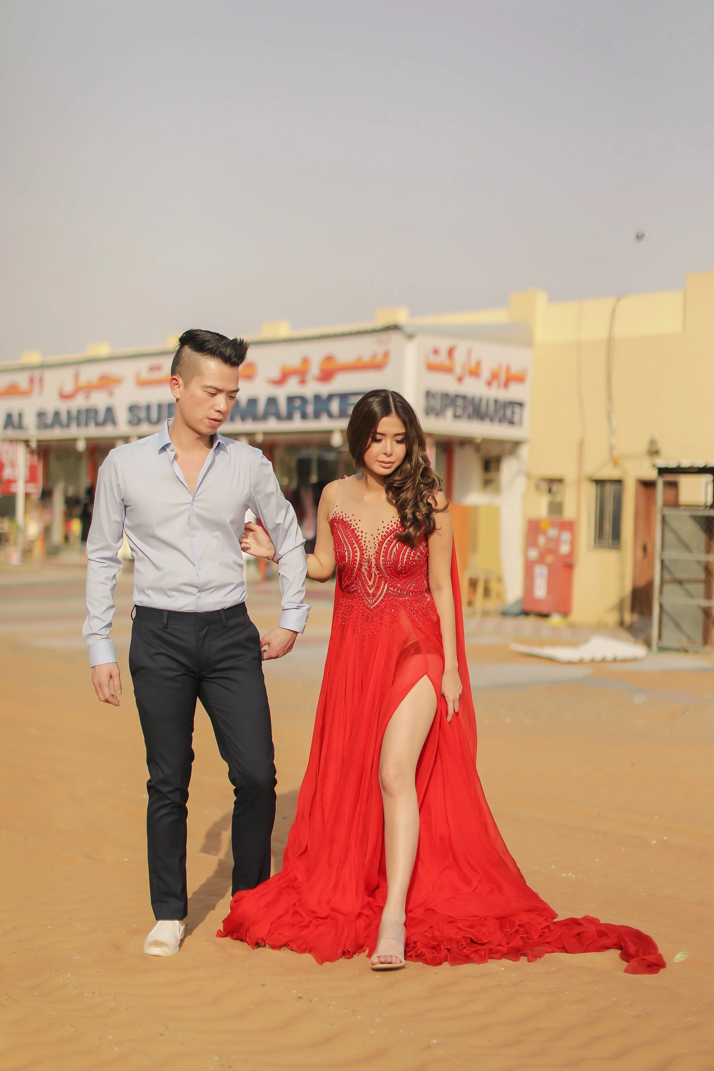 A woman in a red dress with a high slit is walking with a man in a light blue dress shirt and black pants on a sandy surface in front of a market building called Supermarket.
