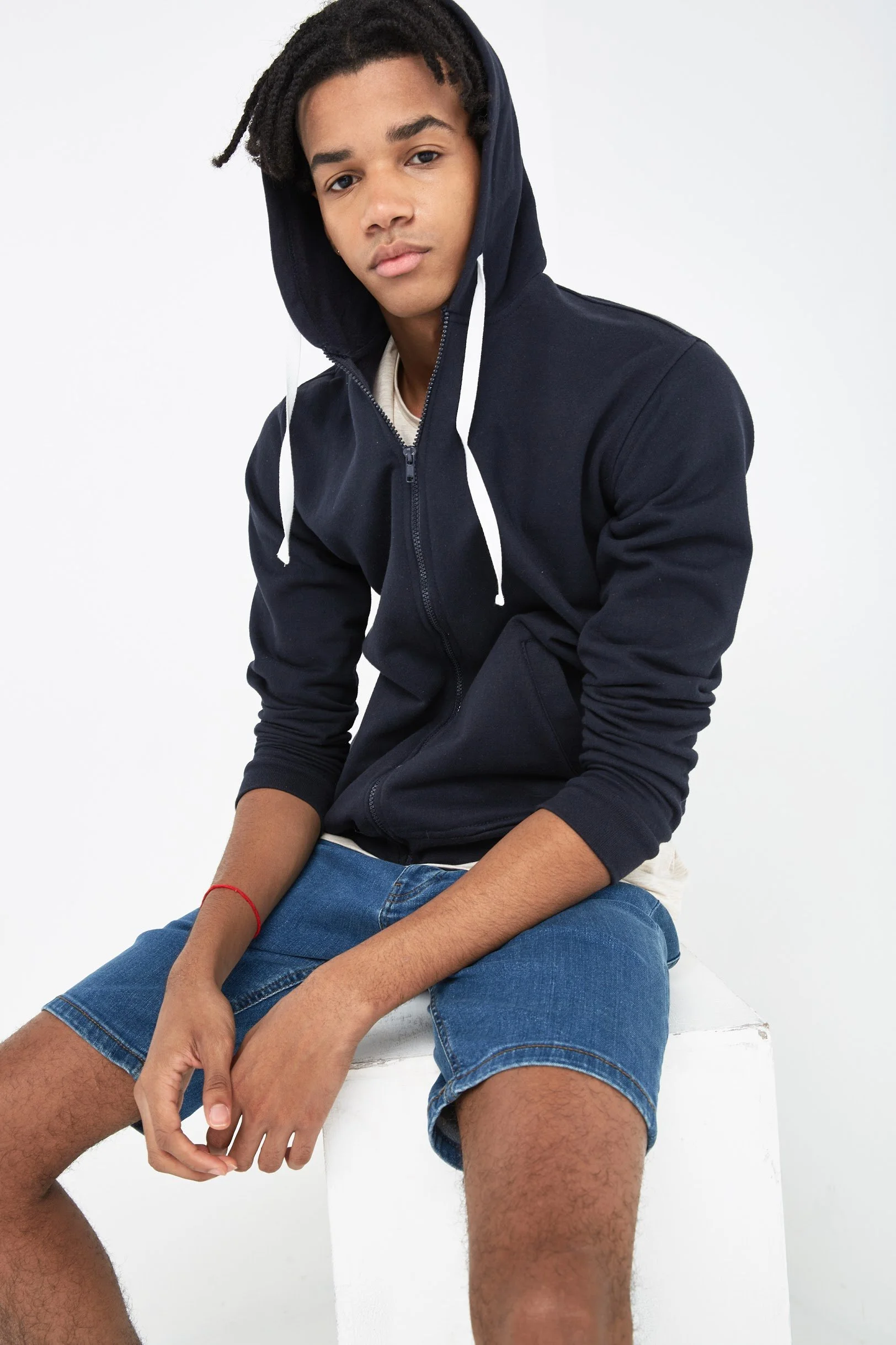 A young man with dark, dreadlocked hair sits on a white block against a plain white background. He is wearing a navy zip-up hoodie with the hood up, a light-colored T-shirt underneath, and blue denim shorts.
