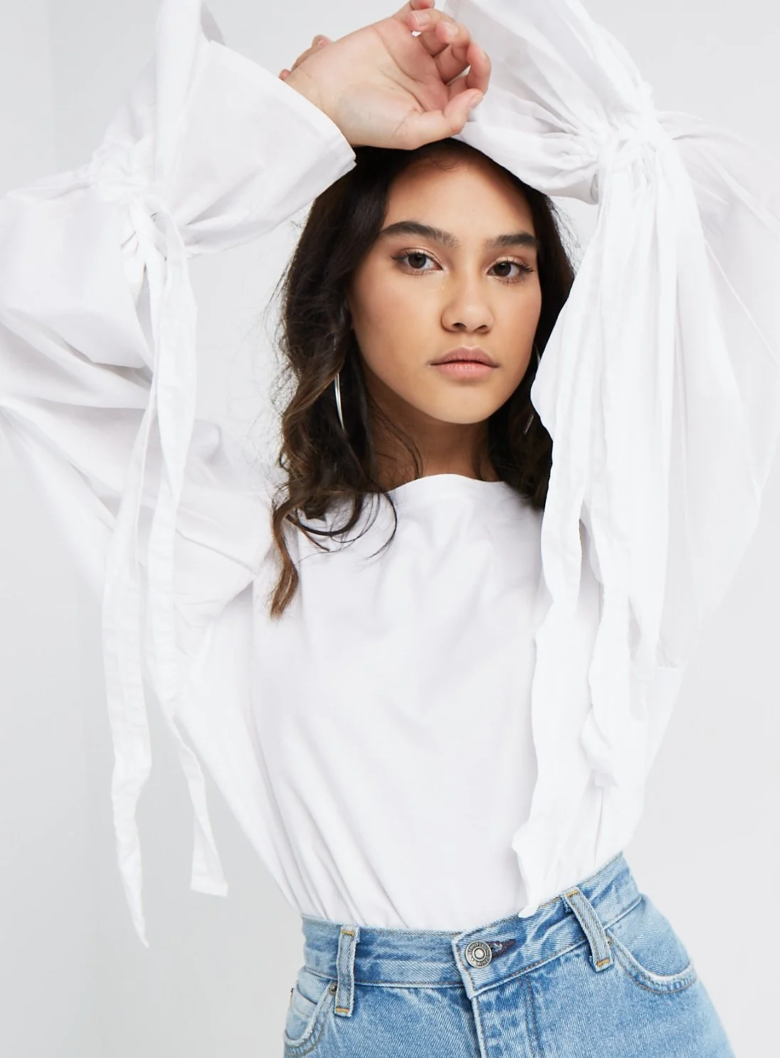 A young woman with wavy dark hair, wearing hoop earrings, a white top, and light blue jeans, posing with her hand above her head against a plain background.