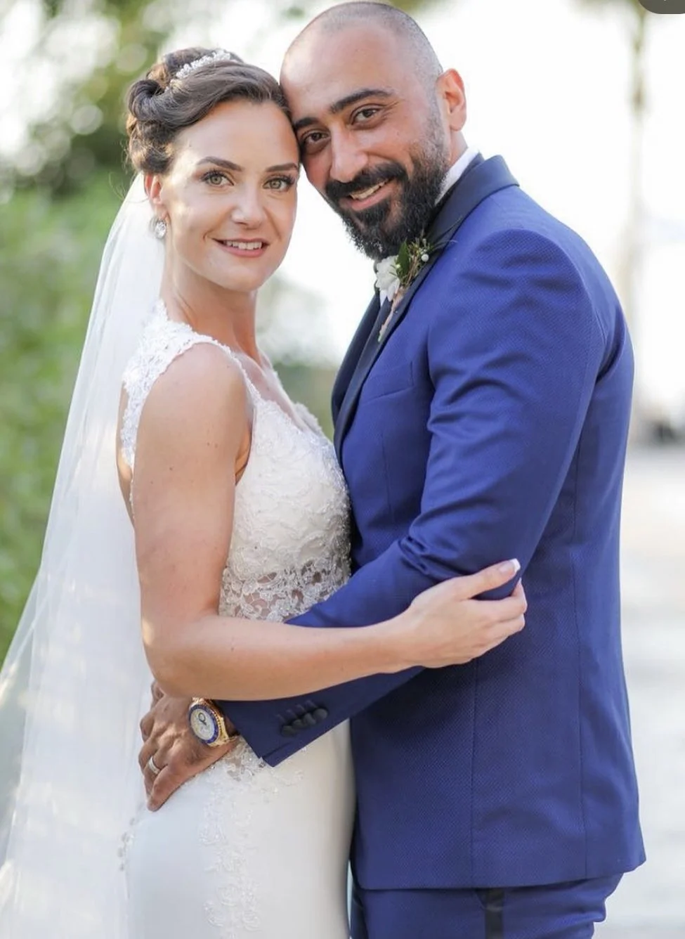 A bride and groom embrace on their wedding day, outdoors, with greenery in the background. The bride wears a white lace wedding dress and veil. The groom wears a blue suit with a white shirt, dark tie, and boutonniere.