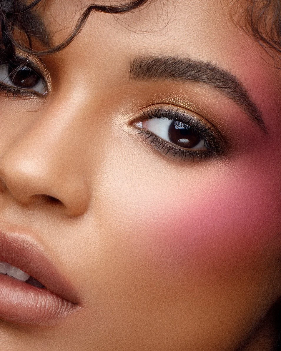 Close-up of a person's face with makeup, featuring bold eye makeup with eyeshadow and long lashes, natural lip color, and pink blush on the cheek.