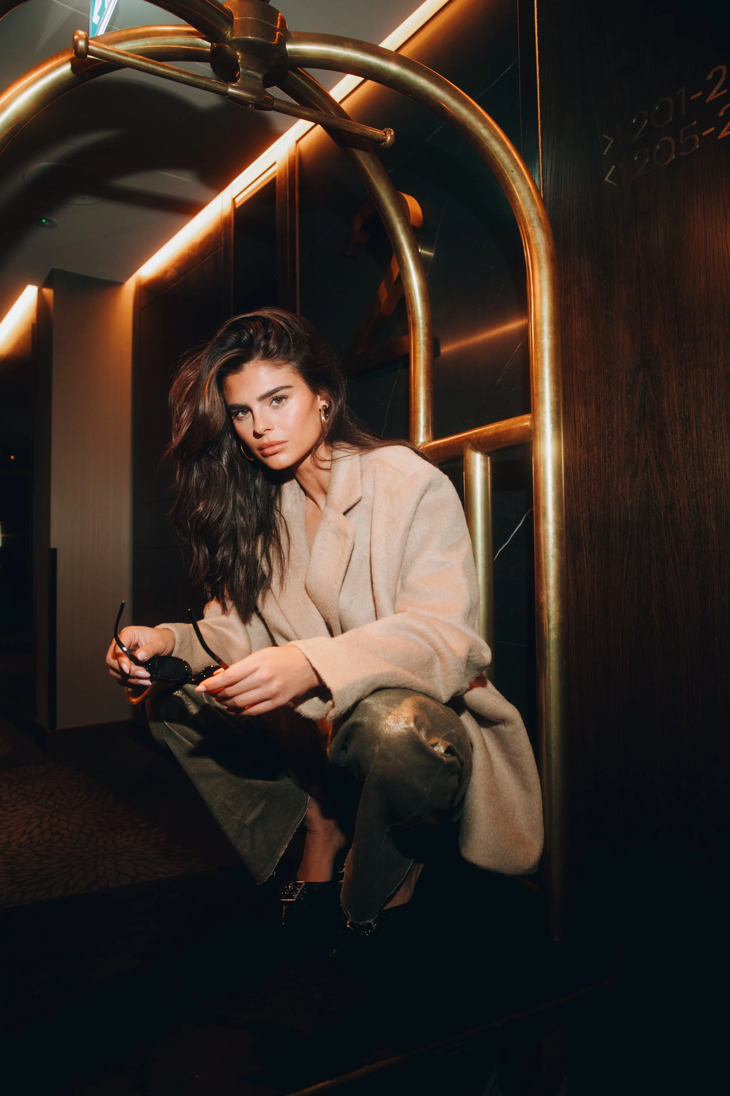 A woman with dark wavy hair, wearing a beige coat and camo pants, squatting on the floor of a dimly lit room with modern decor, holding sunglasses.