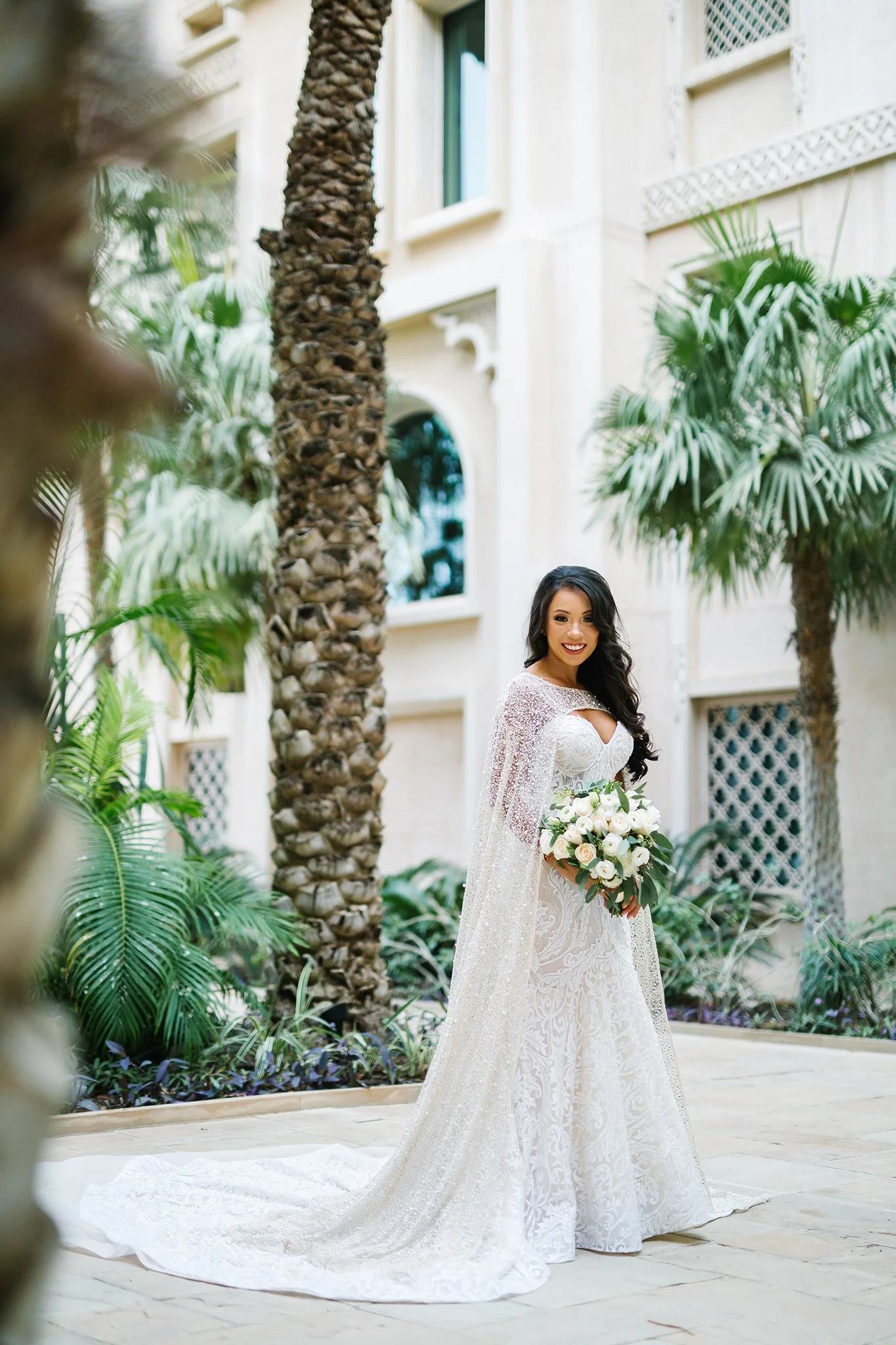 A bride in a white wedding gown holding a bouquet of white flowers, standing outdoors near palm trees and a cream-colored building.