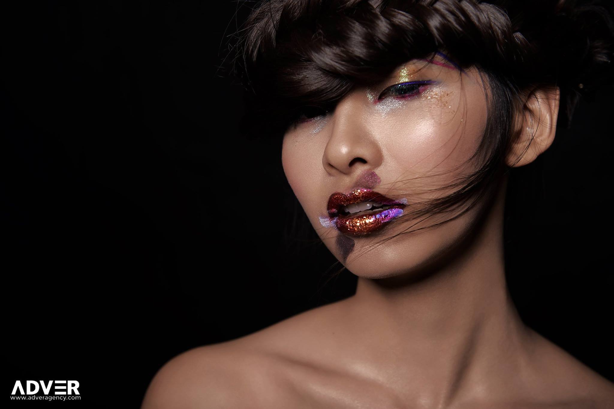 Close-up of a woman with multicolored glitter makeup on her eyes and lips, set against a black background.