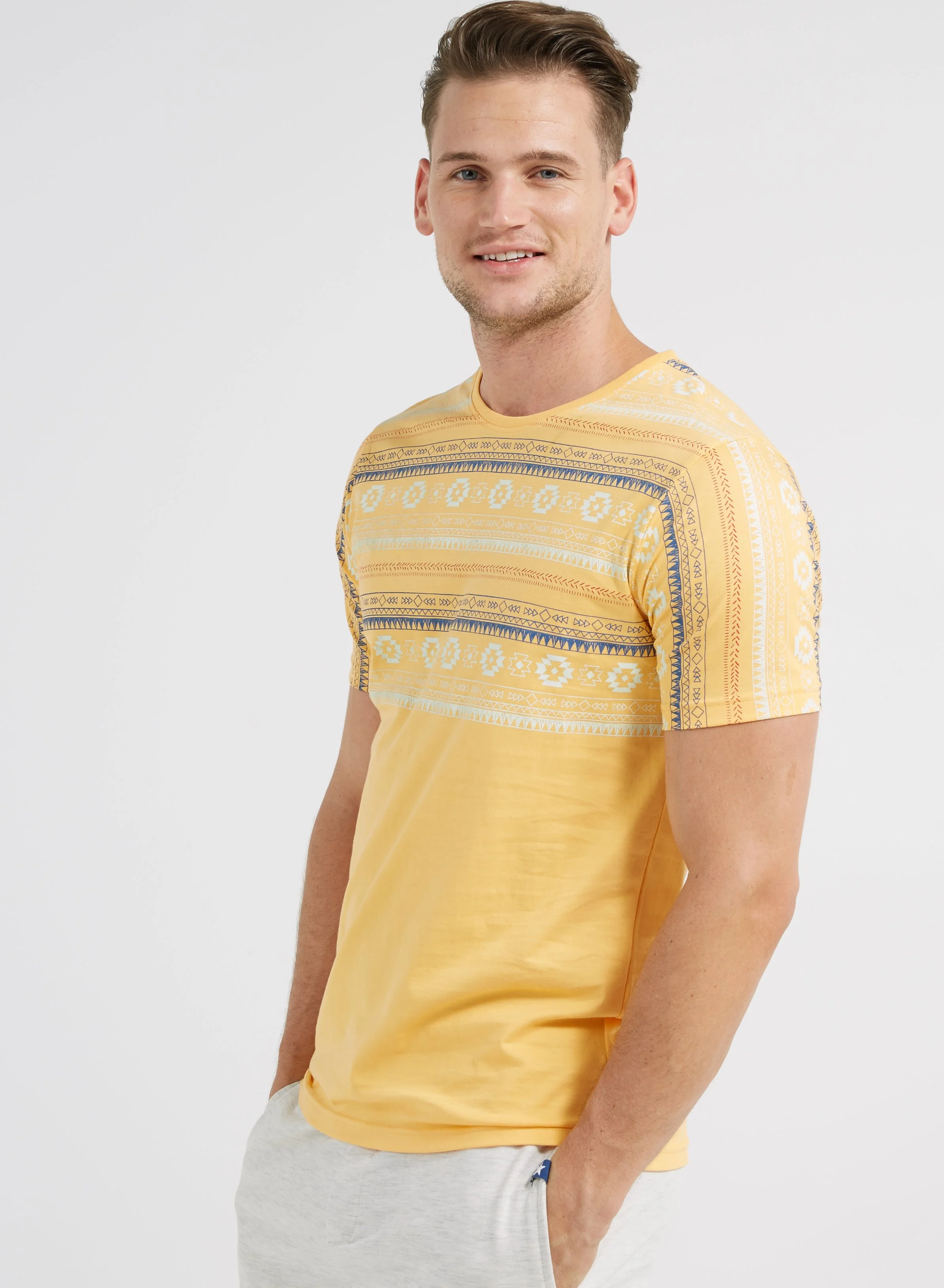 A young man with light skin and short light brown hair, smiling, wearing a yellow T-shirt with a blue and white geometric pattern across the chest and sleeves, and light gray shorts, standing against a plain white background.