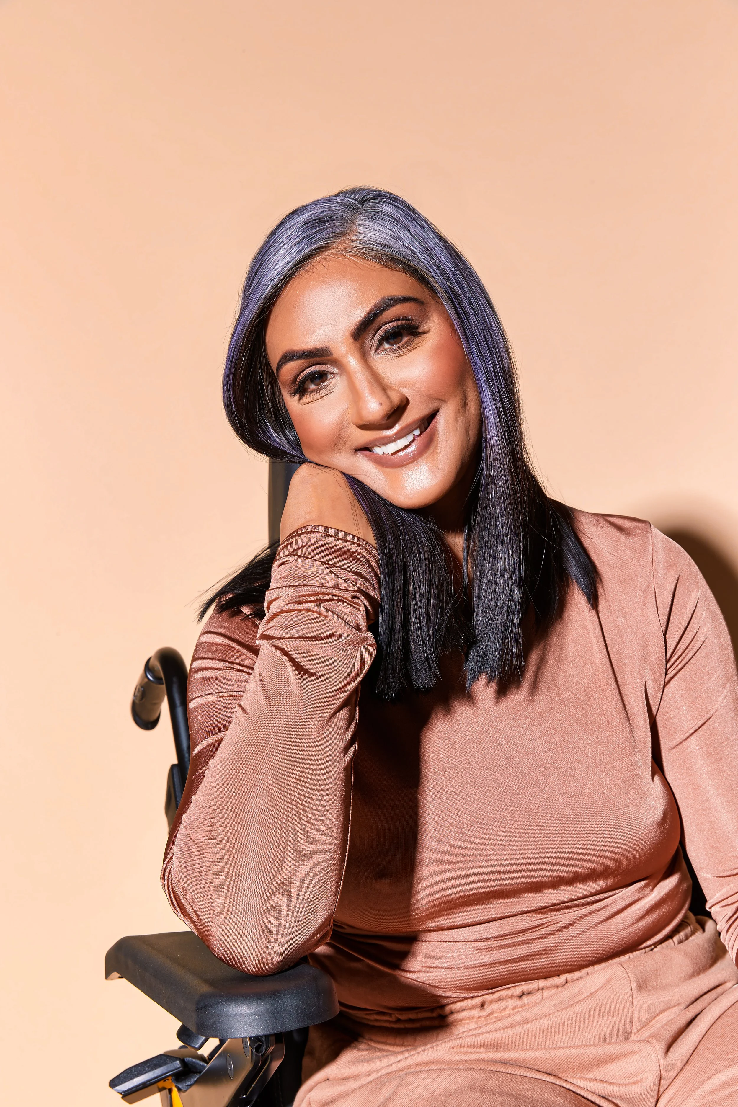 A woman with dark hair and light skin, sitting in a wheelchair, smiling and resting her head on her hand against a beige background.