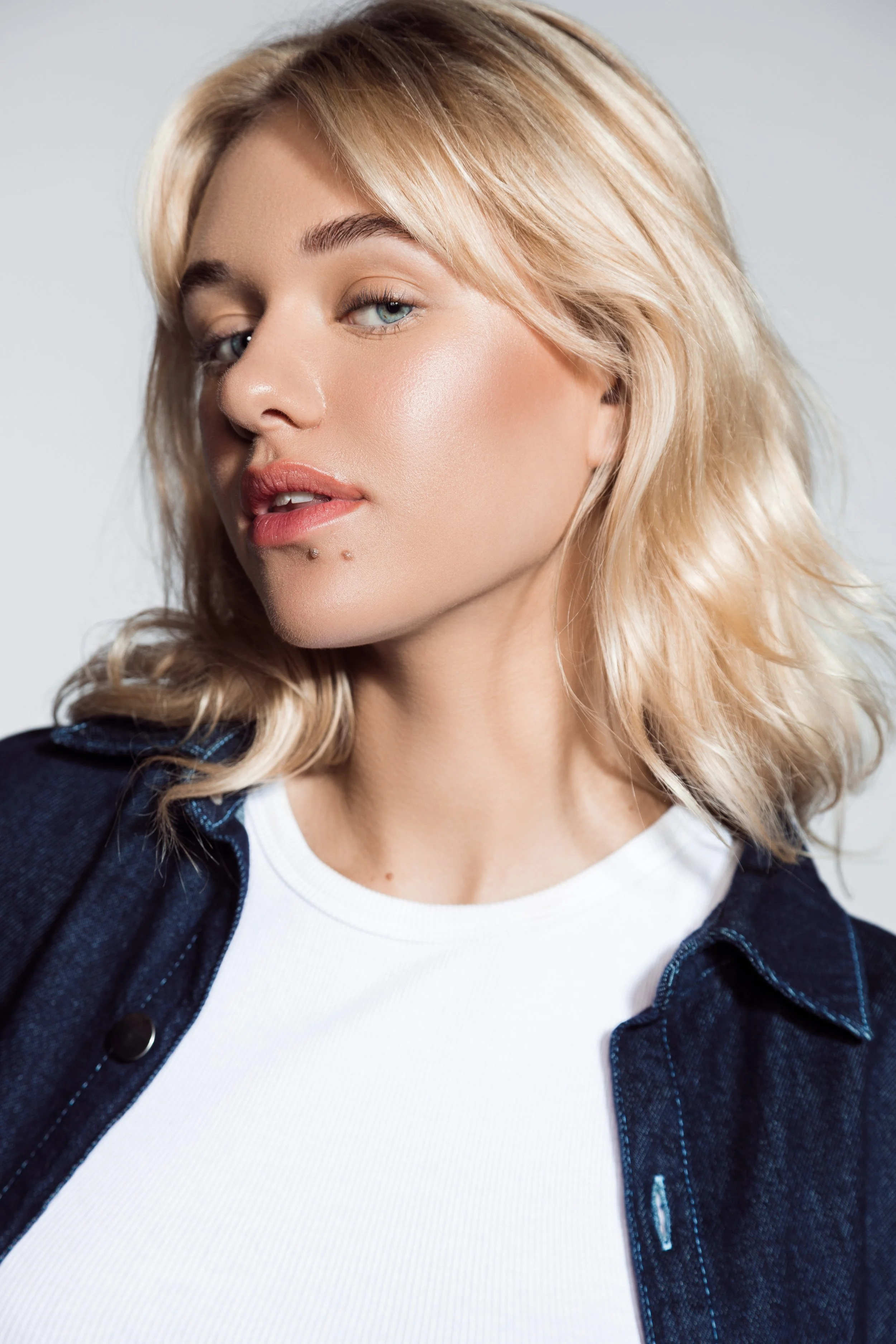 A close-up of a young woman with blonde, wavy hair, fair skin, and blue eyes, wearing a white shirt and a dark denim jacket, against a plain light background.