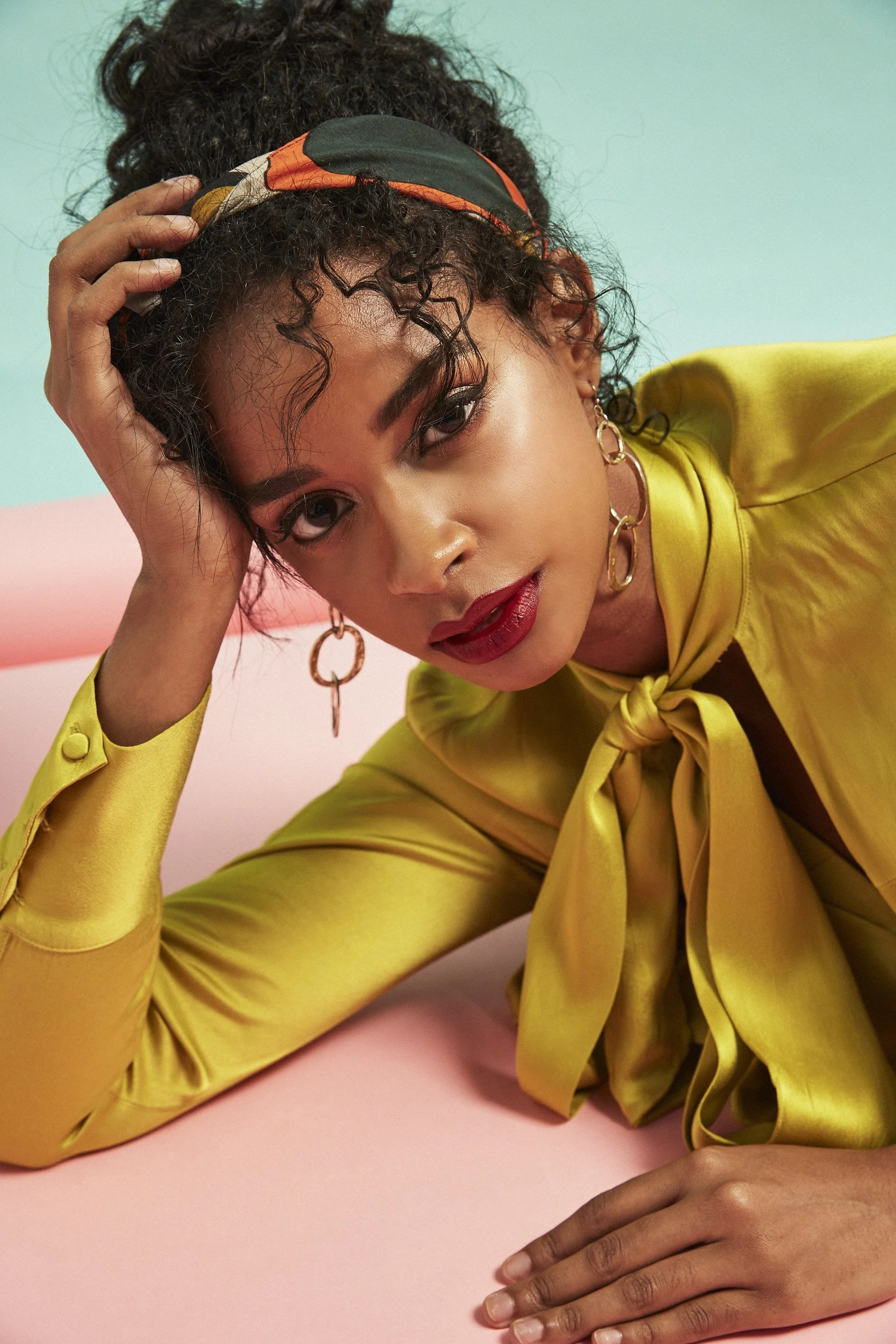 A woman with curly hair, wearing a colorful headband and gold hoop earrings, is lying on a pink surface with a pastel-colored background. She is dressed in a shiny yellow blouse with a tied bow at the collar and has bold makeup with red lipstick.