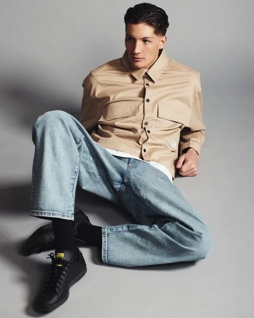 A young man sitting on the floor against a light gray background, wearing a beige button-up shirt, light blue jeans, and black sneakers.