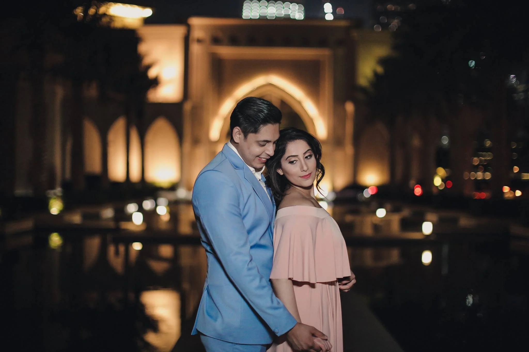 A couple holding hands at night, dressed formally, with an illuminated archway in the background.