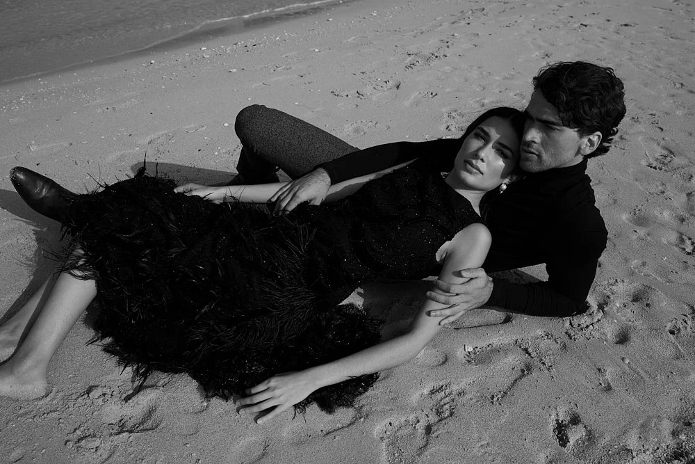 A man and woman lying closely together on a sandy beach, with the woman resting her head on the man's shoulder. The woman is wearing a black outfit, and the man is dressed in dark clothing. The woman’s feet are bare, and she is lying on a black, feat