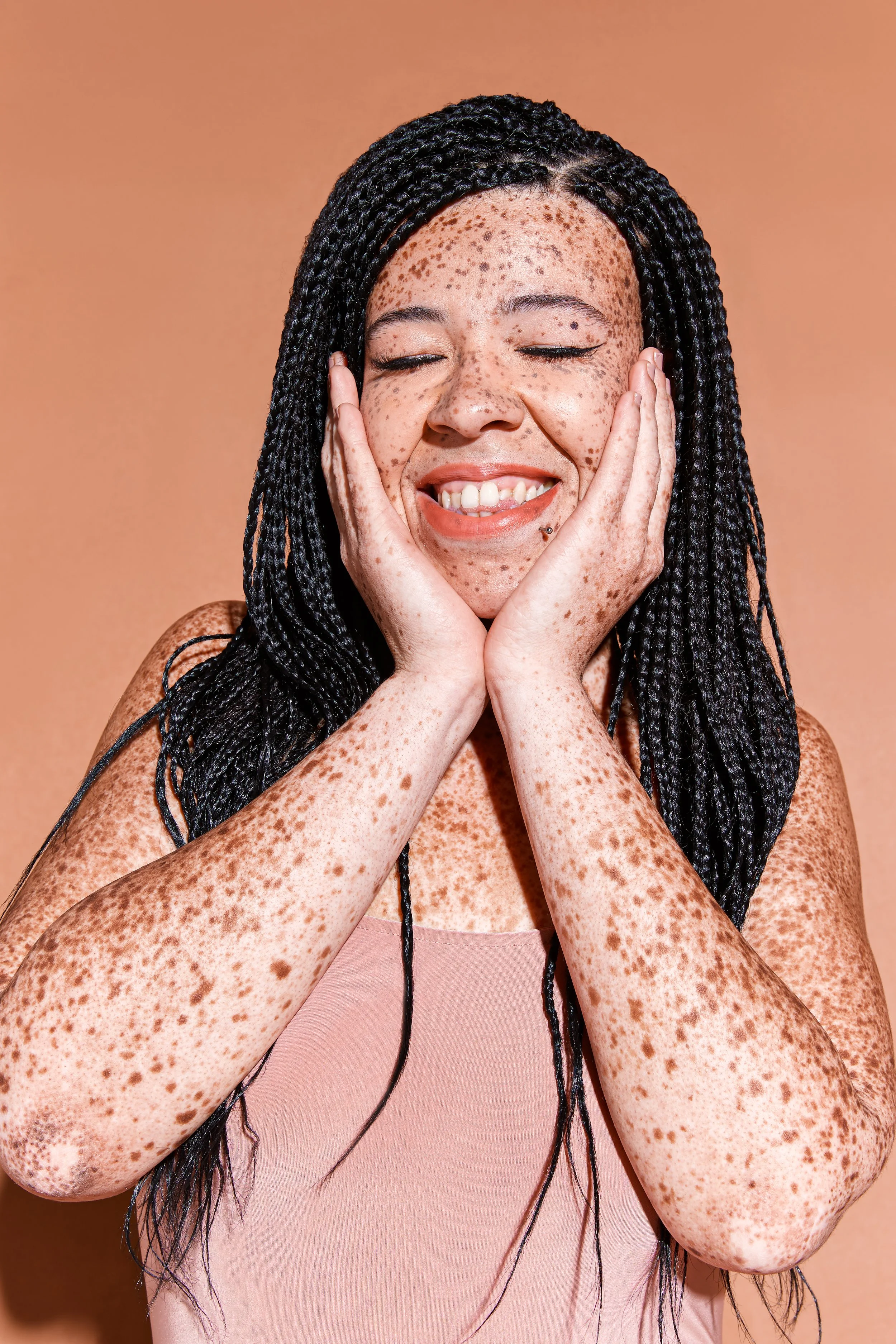 A woman with closed eyes, smiling, with her hands on her cheeks, showing freckles on her face and arms, with long black braided hair, wearing a pink top, against a peach-colored background.