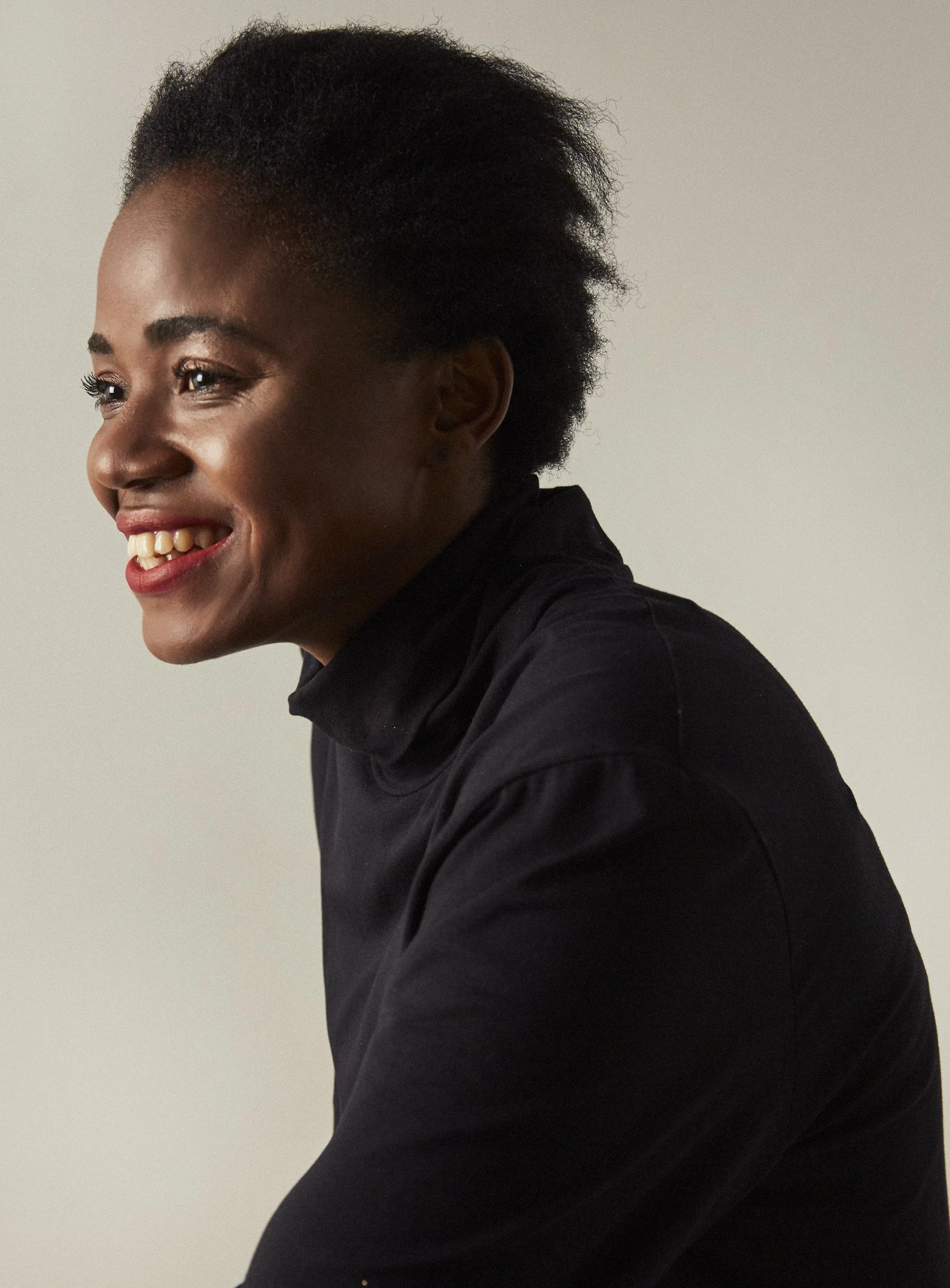 A portrait of a woman smiling, wearing a black turtleneck and sitting against a plain, light-colored background.
