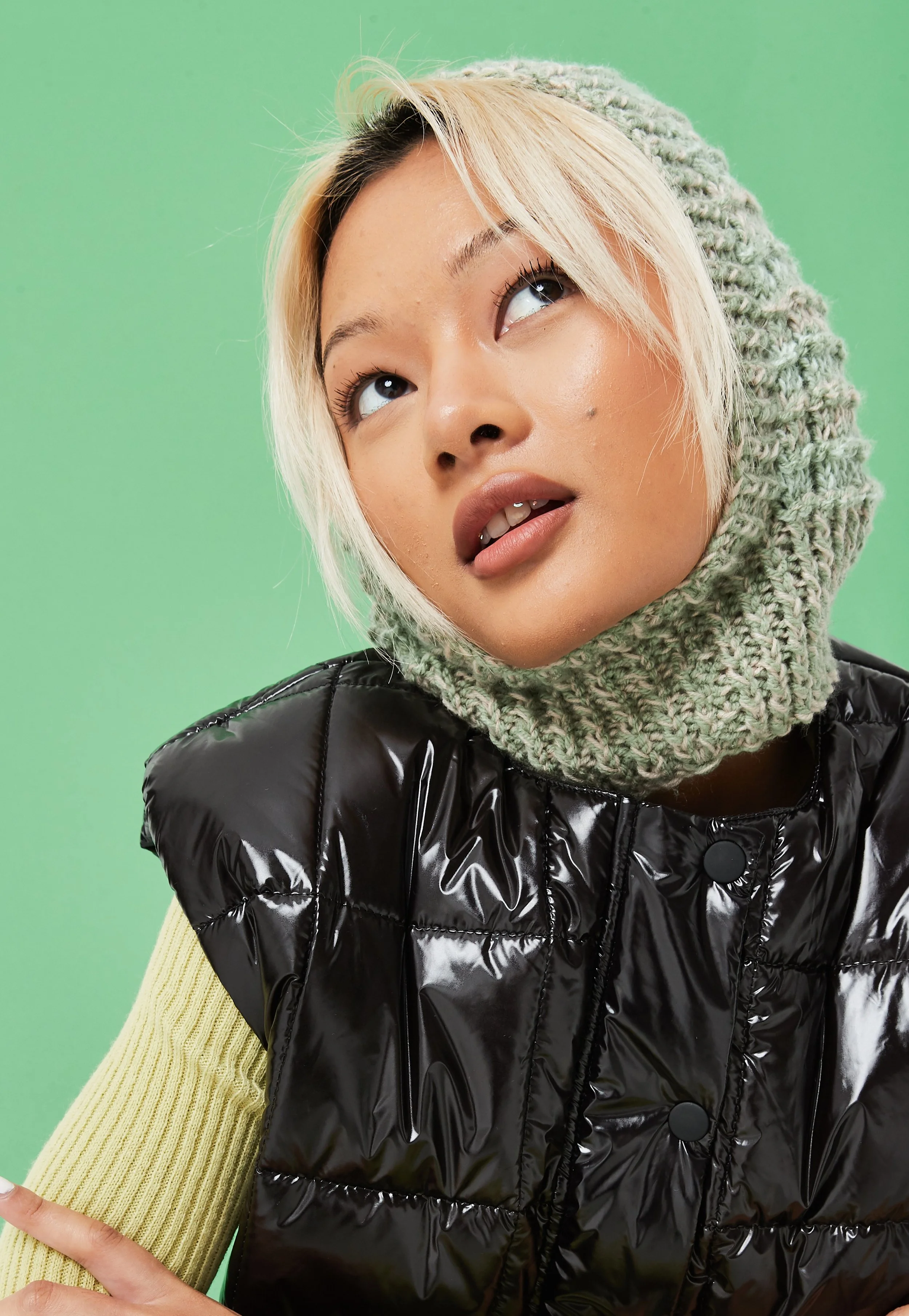 A young woman with blonde hair and makeup, wearing a green knit hat and a black puffer vest, looking upwards against a green background.