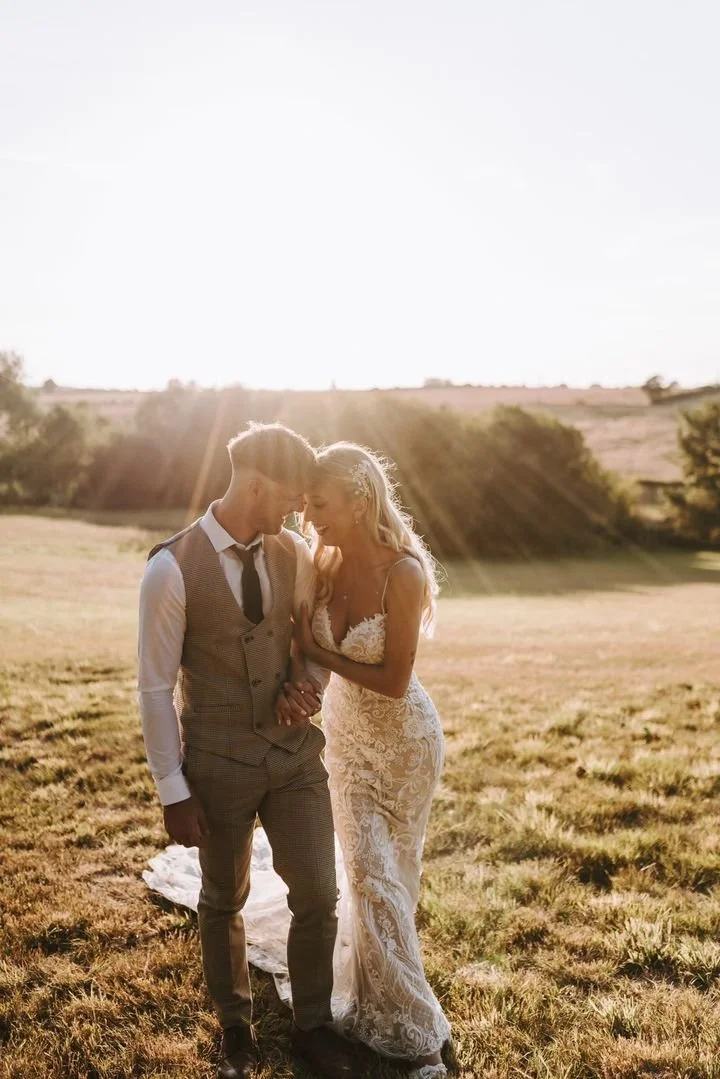 A bride and groom holding hands and gazing at each other in a field during sunset.