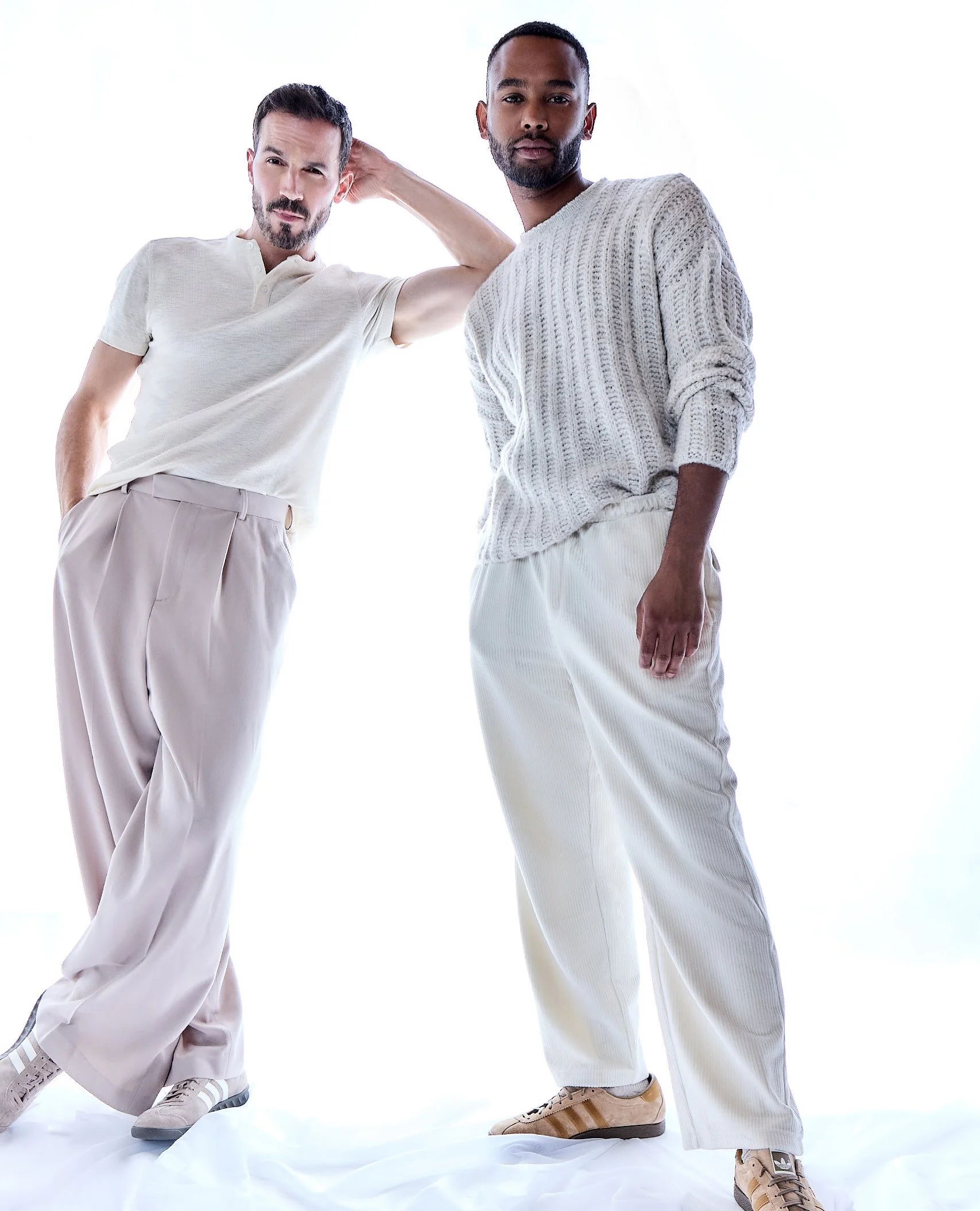 Two men standing against a white background, dressed in neutral-colored casual clothing.