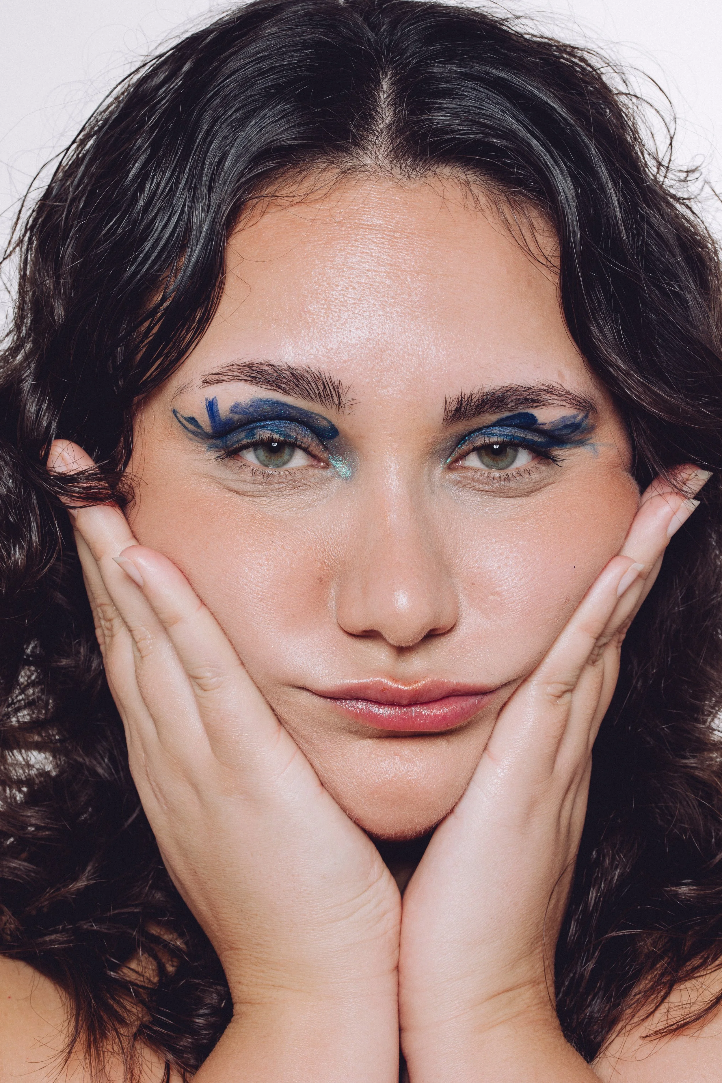 A woman with dark wavy hair and makeup featuring bold blue and black eyeshadow and glittering accents around her eyes, holding her face with both hands.