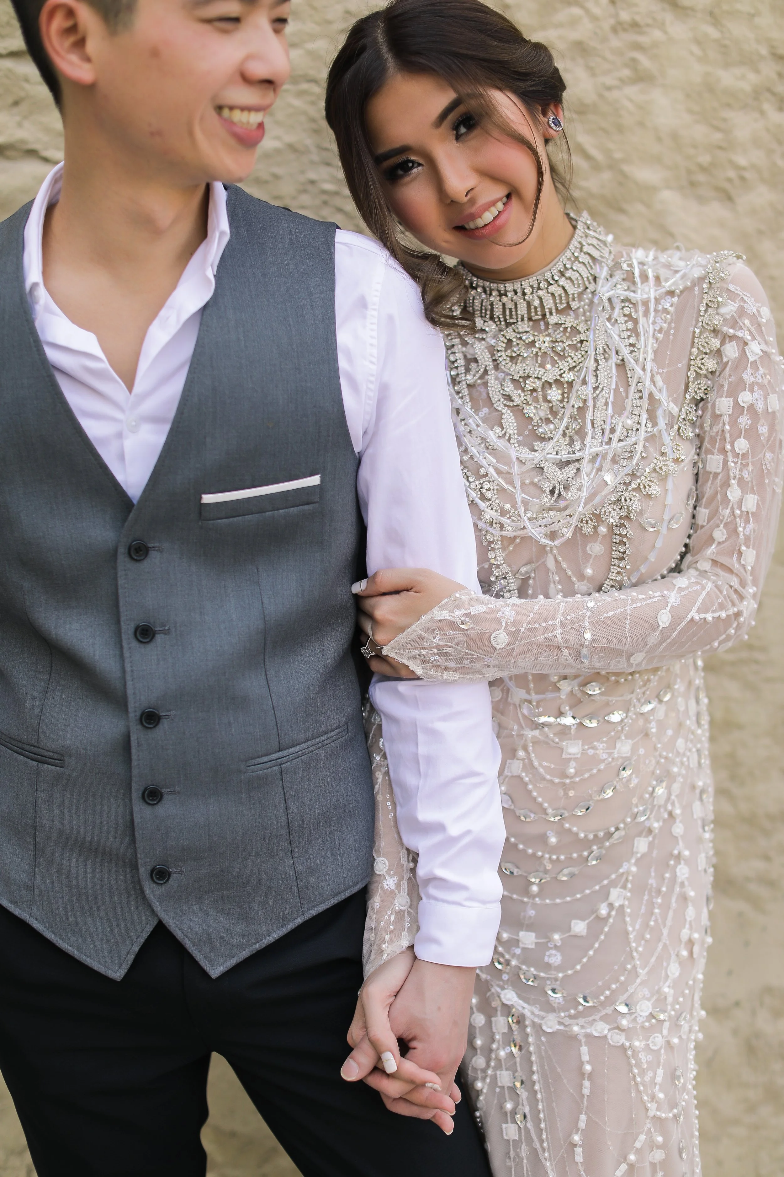 A couple holding hands, with the woman wearing a beaded wedding gown and the man in a gray vest and white shirt, standing against a beige stone wall.