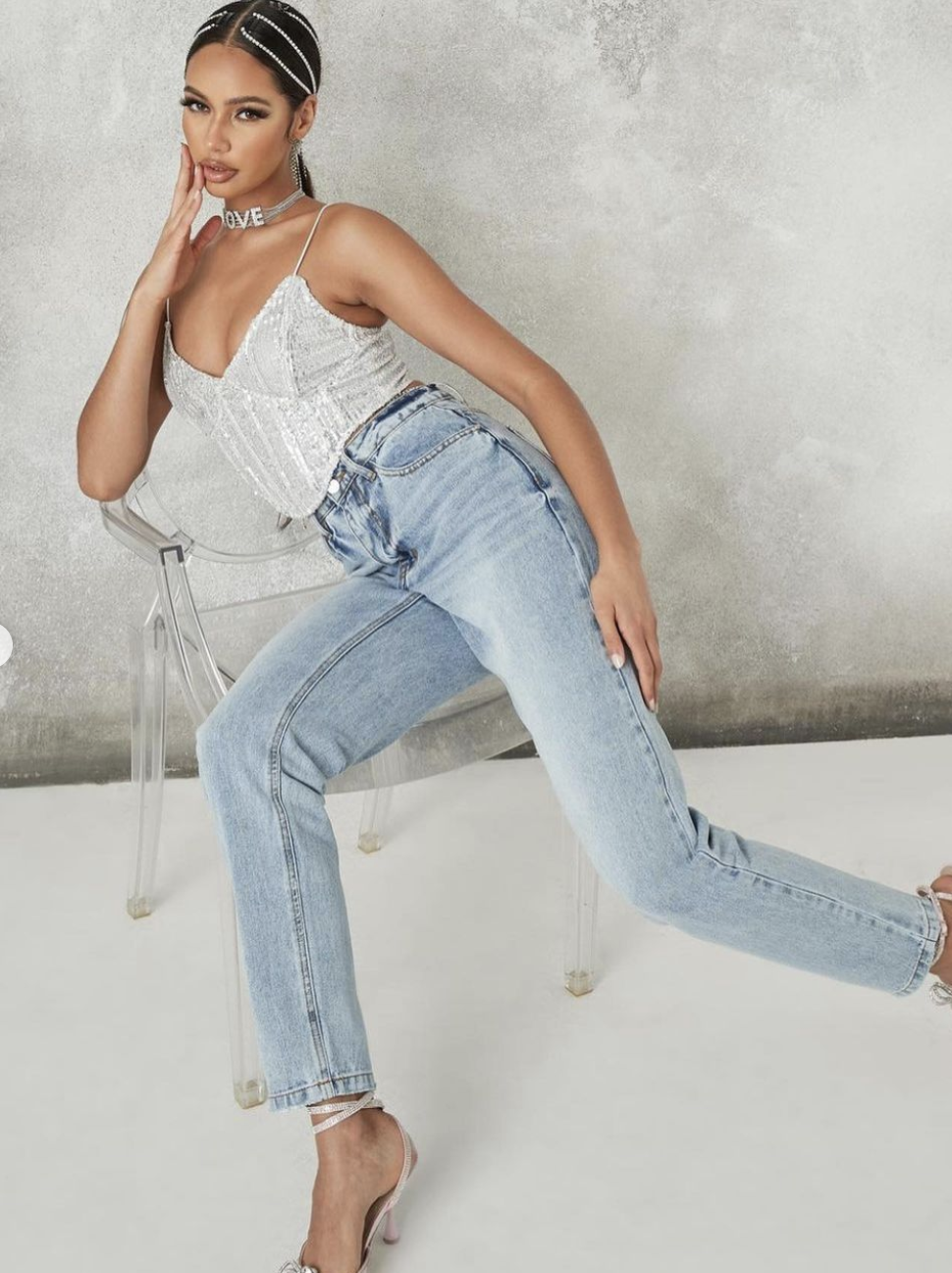 A woman with dark hair styled with pearl hair accessories poses sitting on a clear chair against a plain background. She wears a sparkly silver top, light blue jeans, and matching high heels, with a necklace that says 'LOVE'.