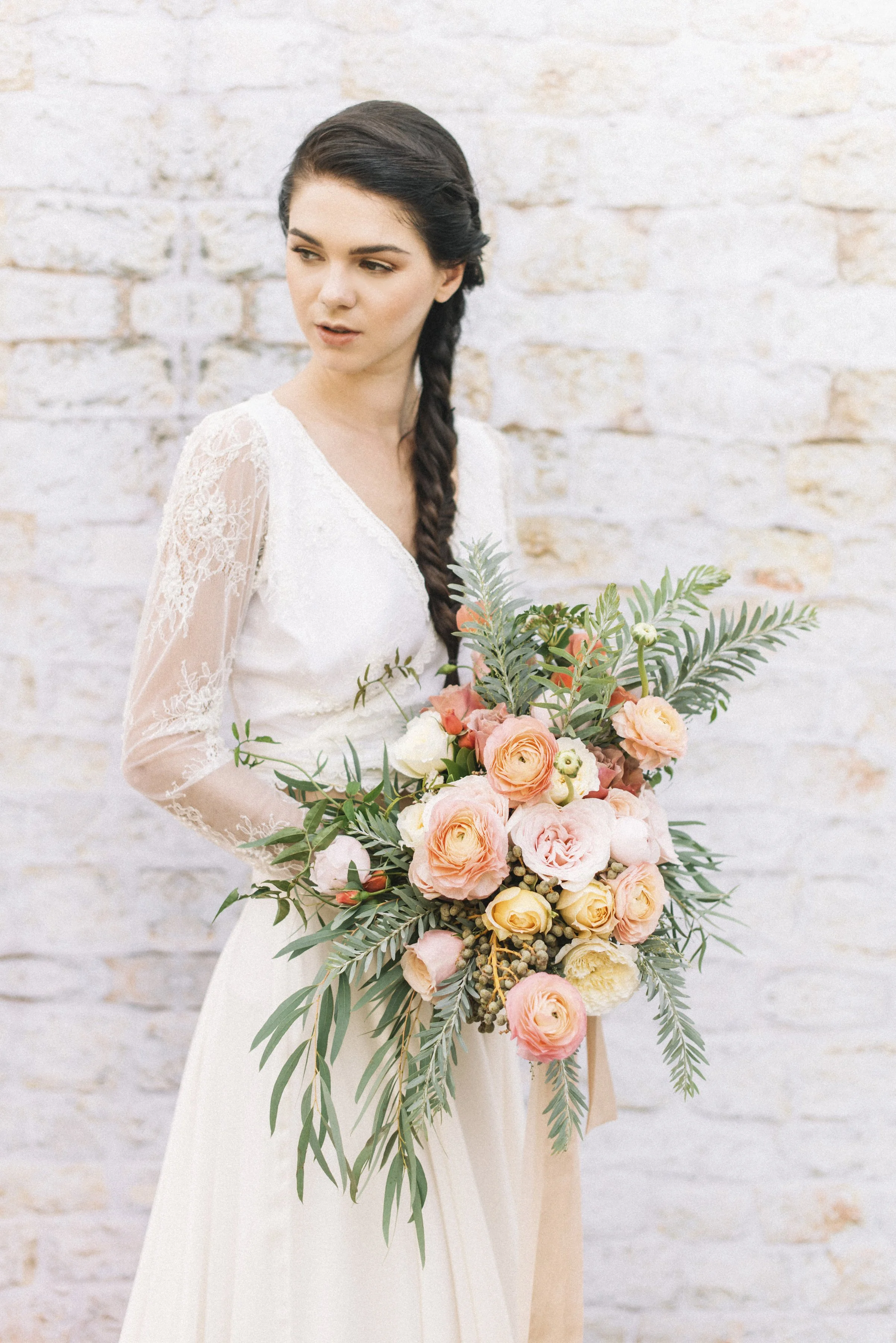 Young woman in a white lace dress holding a bouquet with pink and cream roses, greenery, and eucalyptus leaves, standing against a white brick wall.