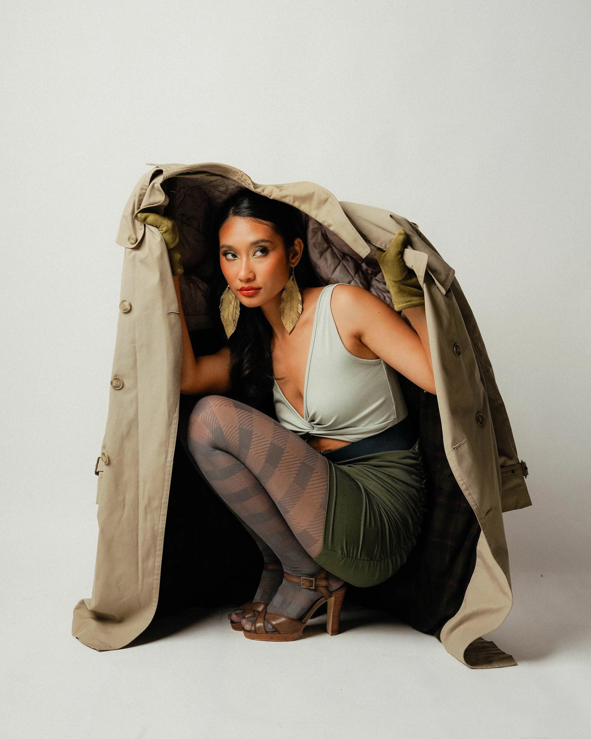 A woman crouching inside a beige trench coat with a plain white background.