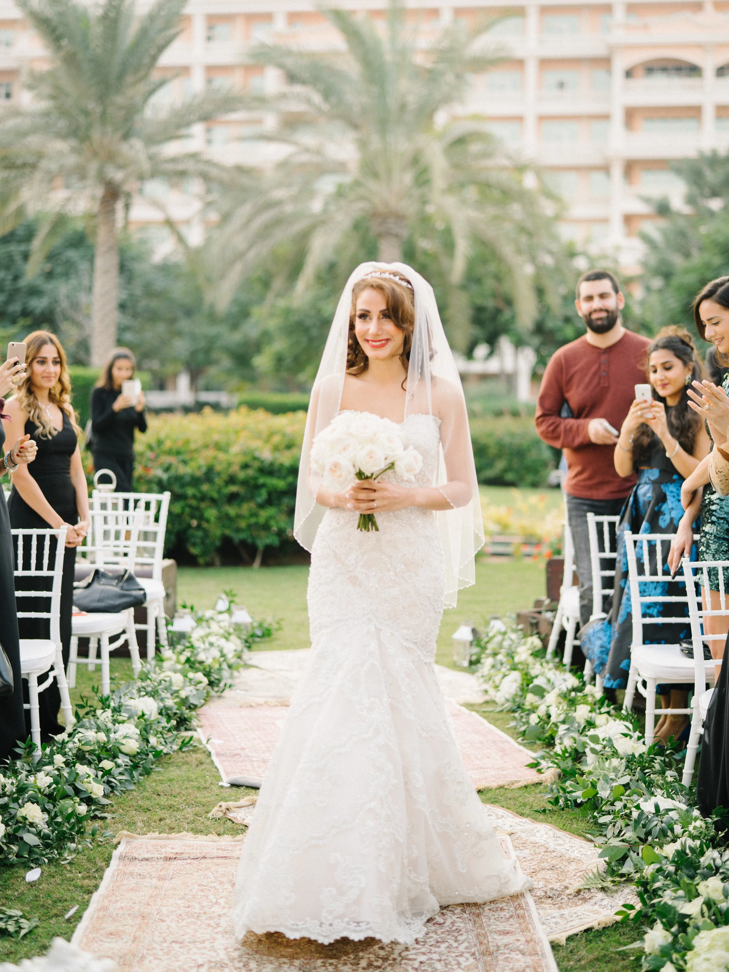 A bride in a white lace wedding dress and veil holding a bouquet of white flowers, smiling during her outdoor wedding ceremony, while guests take photos and watch.