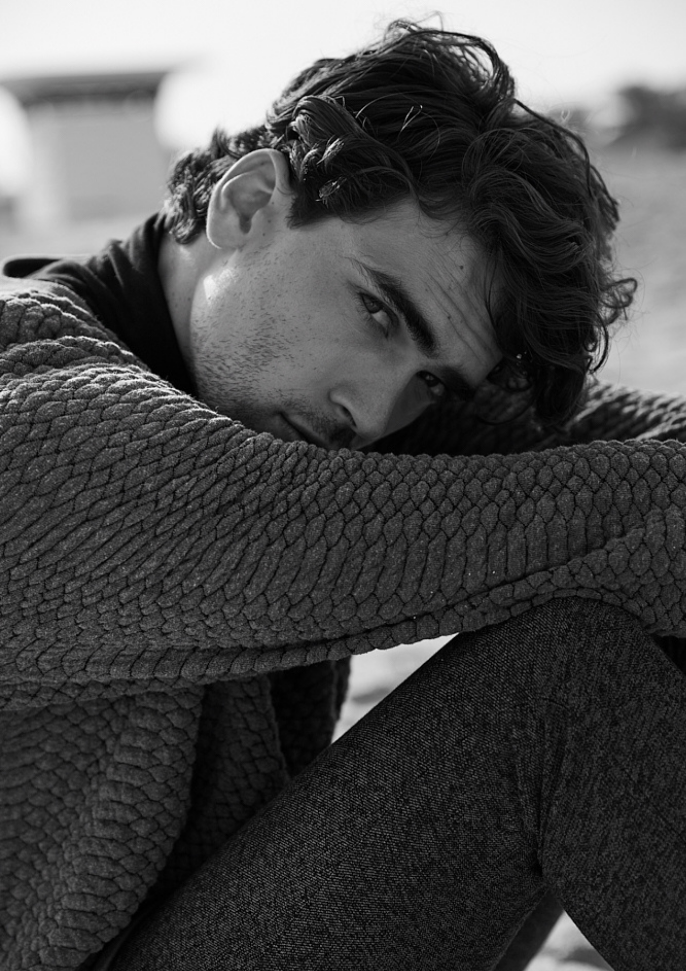 Black and white photo of a young man with curly hair resting his head on his knee, looking at the camera with an intense gaze, outdoors.