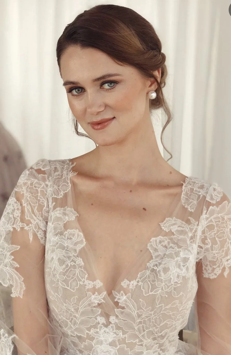 A woman with styled brown hair, wearing pearl earrings and a lace dress, looking at the camera with a slight smile, in front of a light-colored curtain.
