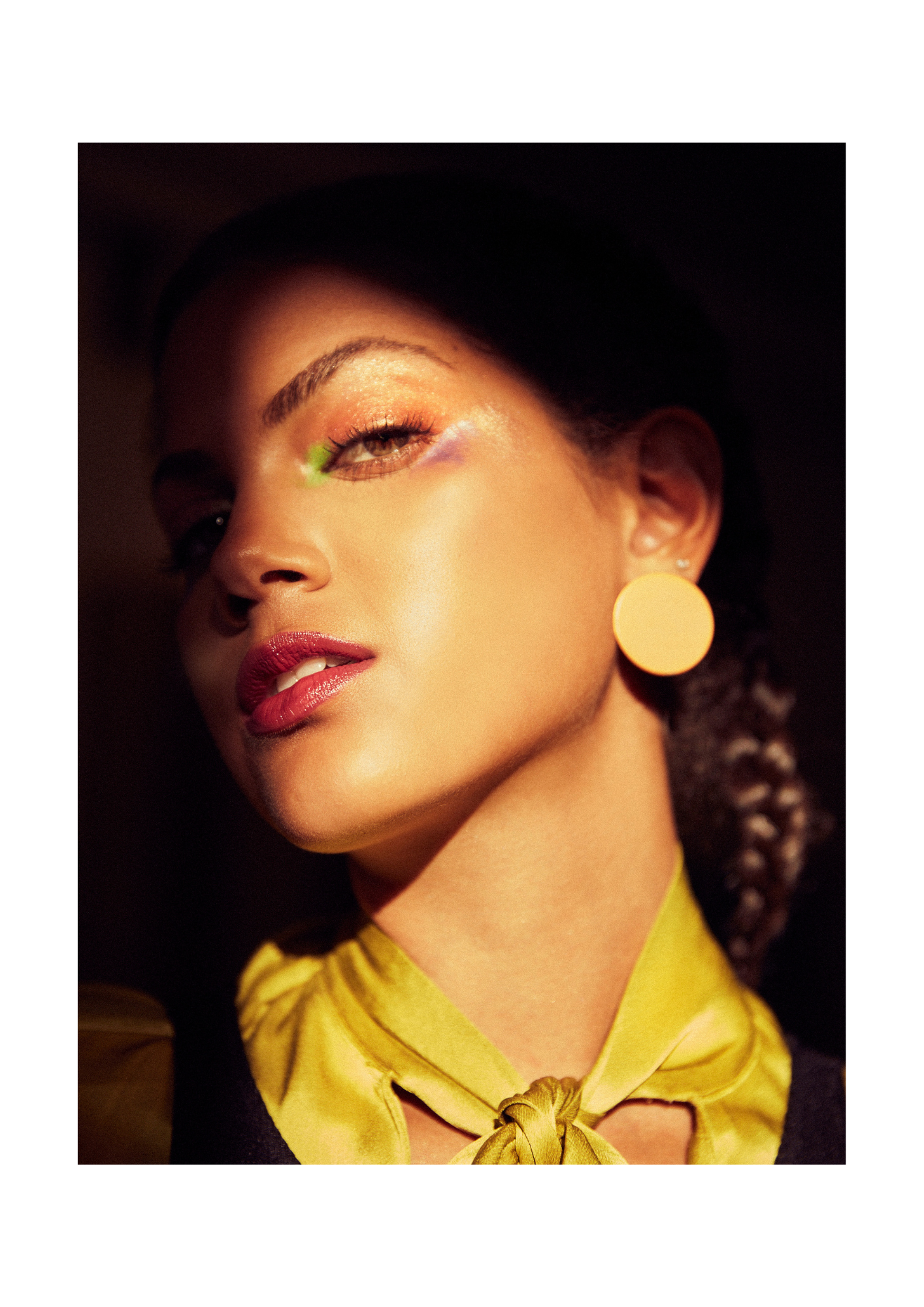 Close-up portrait of a woman with bold makeup, wearing large yellow circular earrings, a yellow scarf tied at her neck, with a confident expression and dark background.
