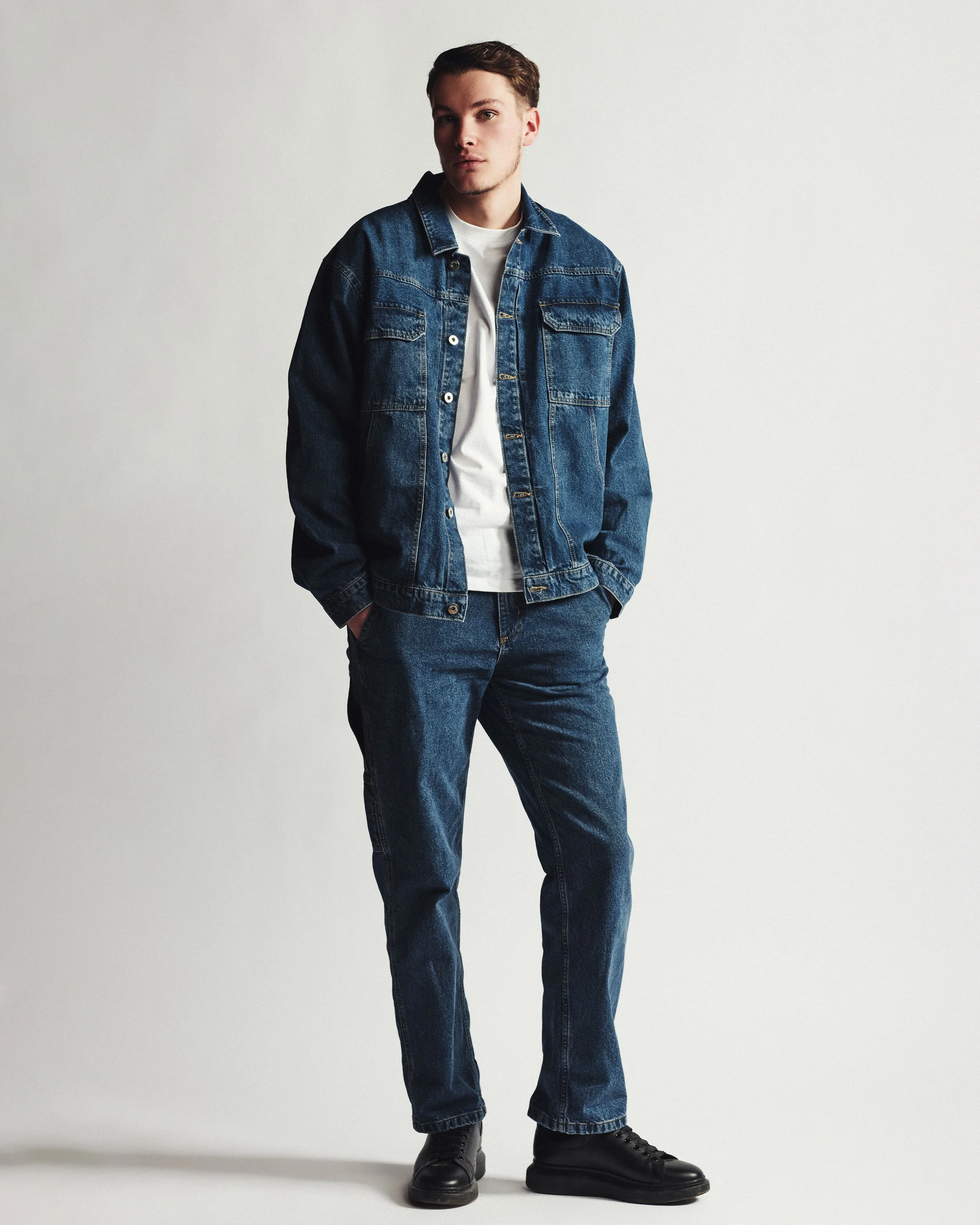A young man wearing a denim jacket and jeans, white t-shirt, black shoes, standing against a plain background.
