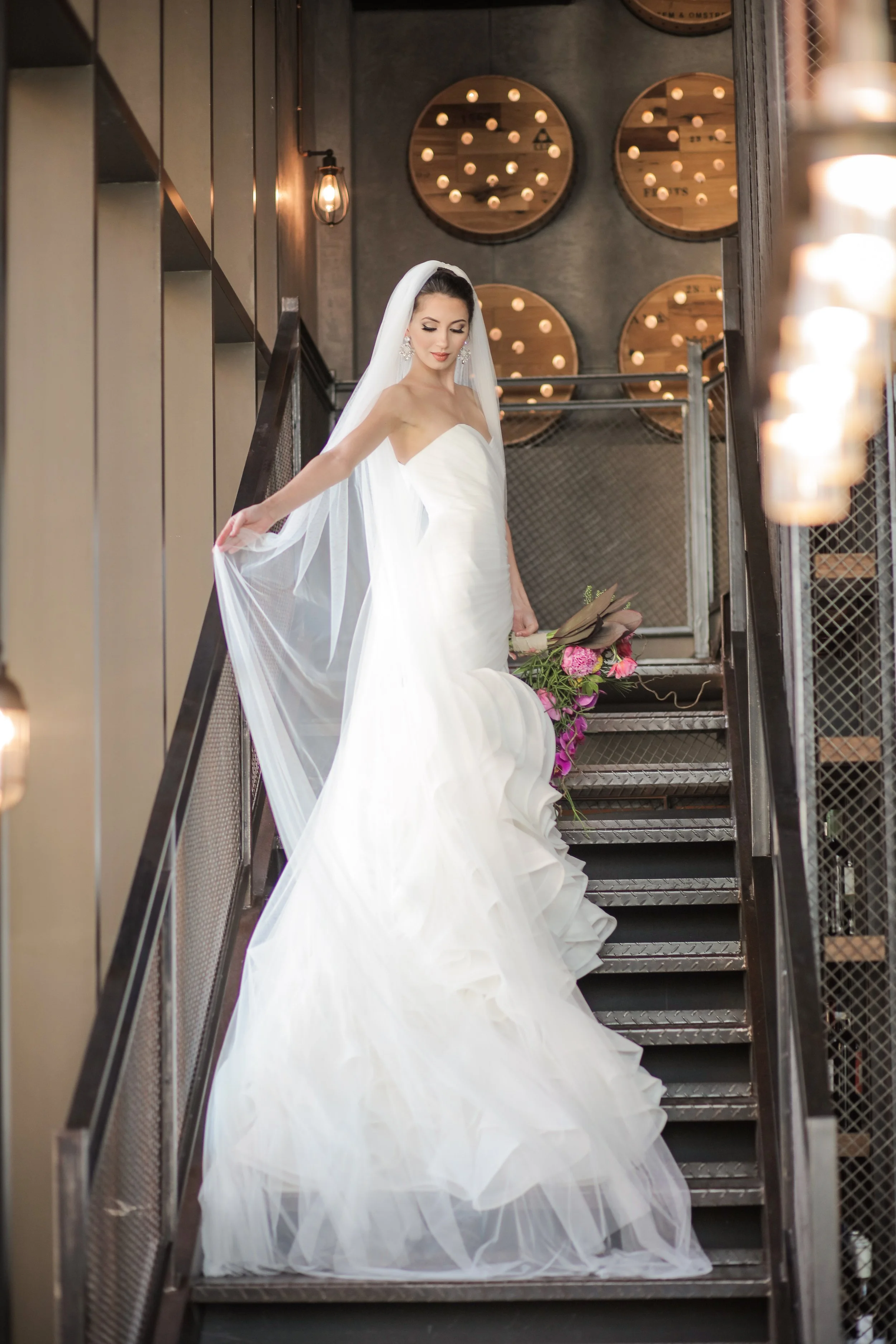 A bride wearing a white strapless wedding gown with a long flowing veil, holding a bouquet of pink and purple flowers, standing on metal stairs inside a modern industrial-style building.