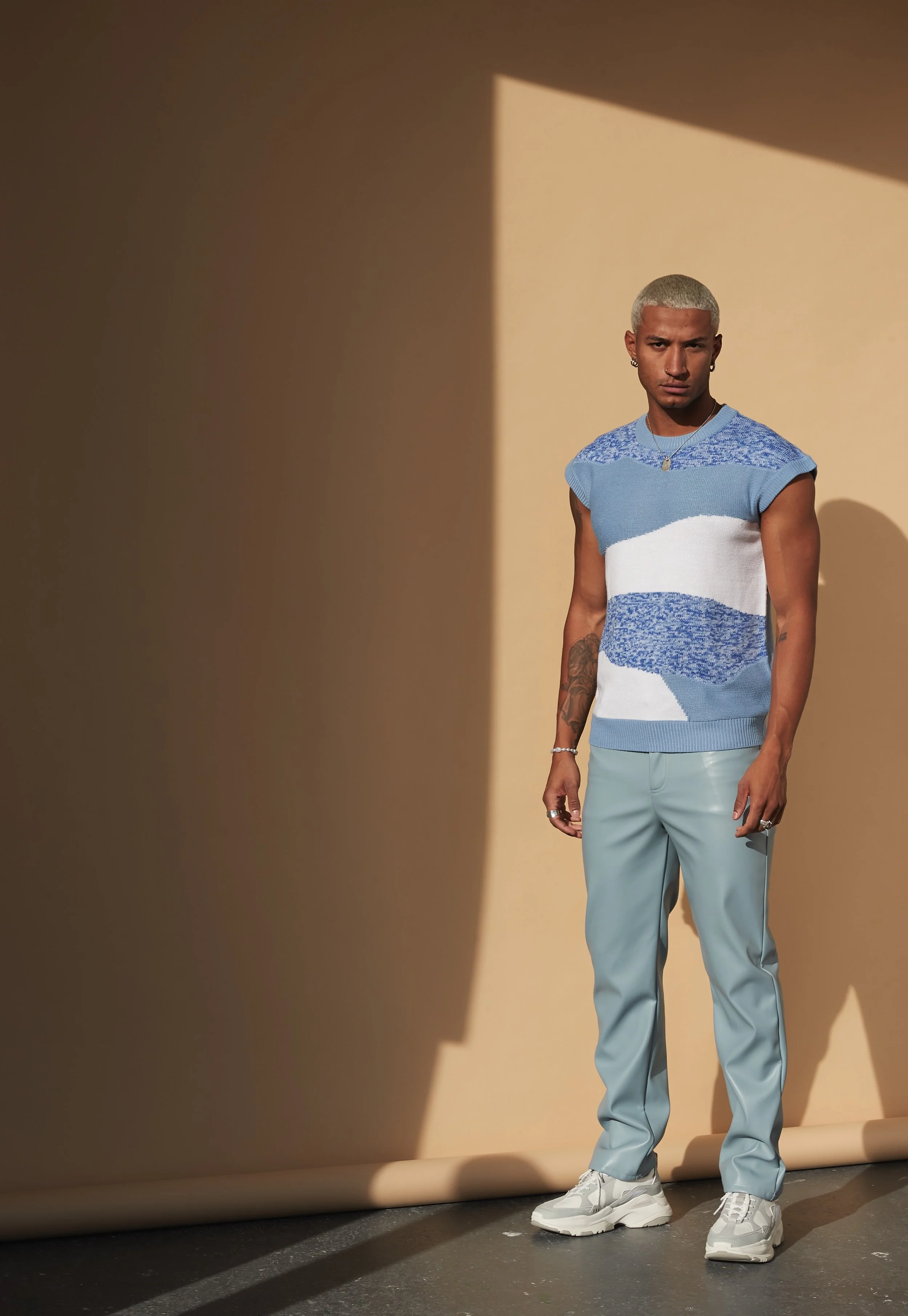 A young man with bleach-blonde hair stands in front of a beige backdrop, wearing a blue and white sleeveless sweater, light blue pants, and white sneakers. The lighting creates a shadow on the backdrop.