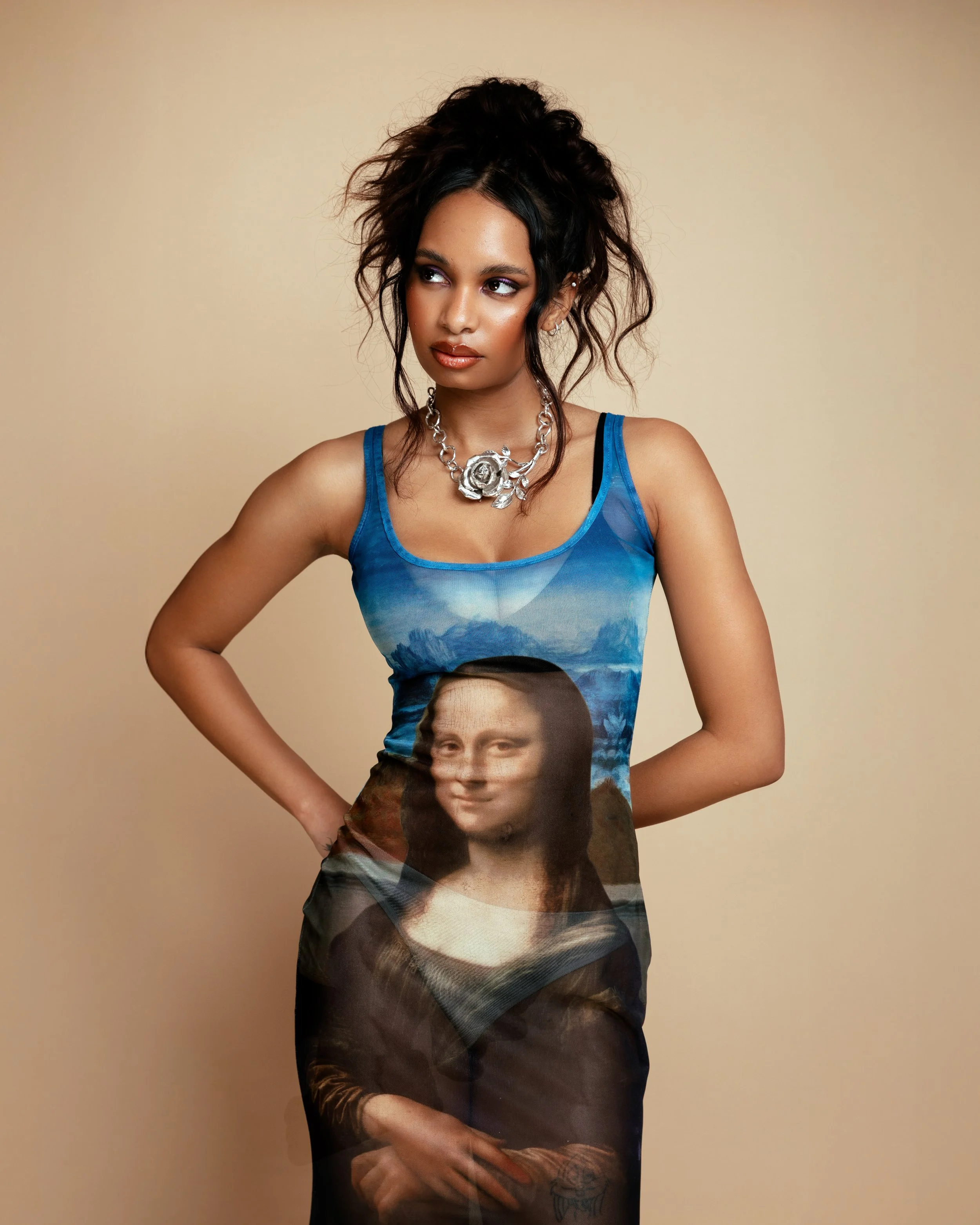 A woman with dark hair in a messy updo, wearing a blue tank top with the Mona Lisa painting printed on it, standing against a beige background.