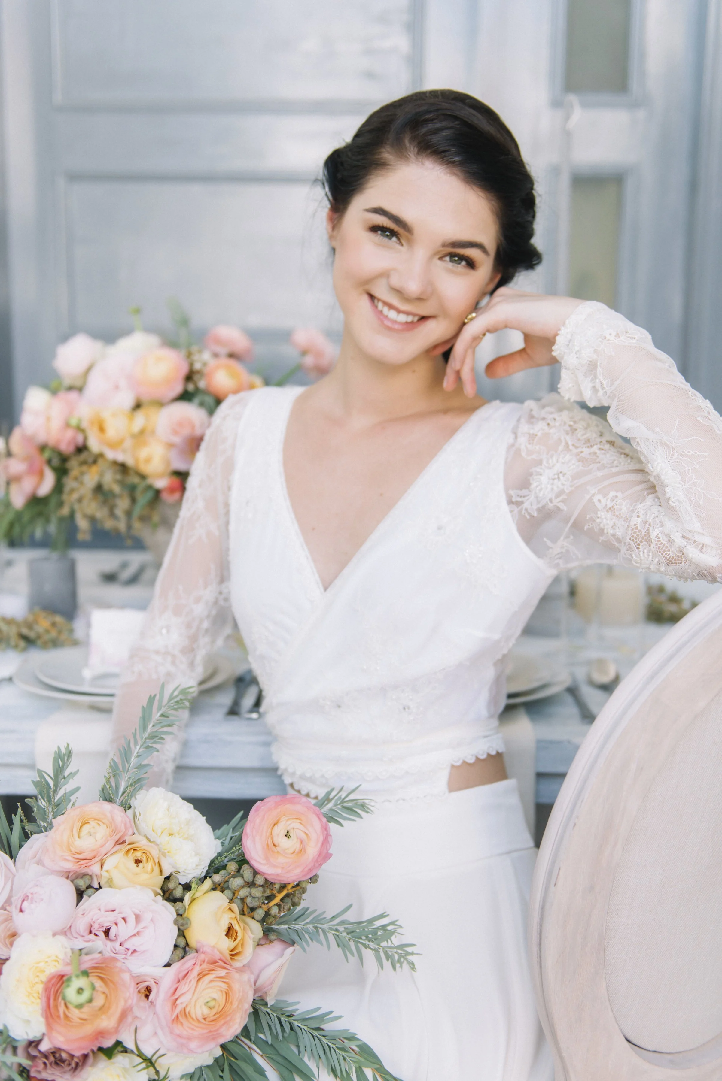 A bride smiling, wearing a white lace wedding dress, sitting at a decorated table with a bouquet of pink, yellow, and cream flowers, with a background of a rustic wooden wall and large window.