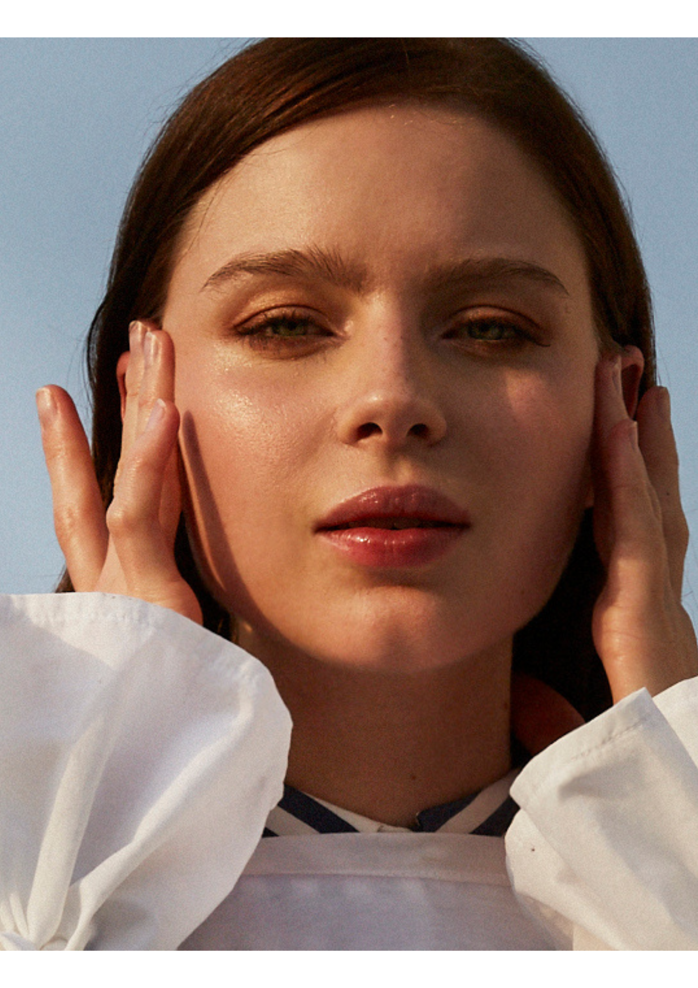 Close-up of a woman with brown hair, holding her face with both hands, against a clear blue sky.
