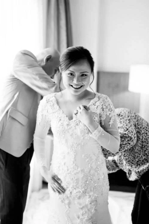 A woman in a wedding dress smiling and making a fist in celebration, with two people behind her adjusting her dress.